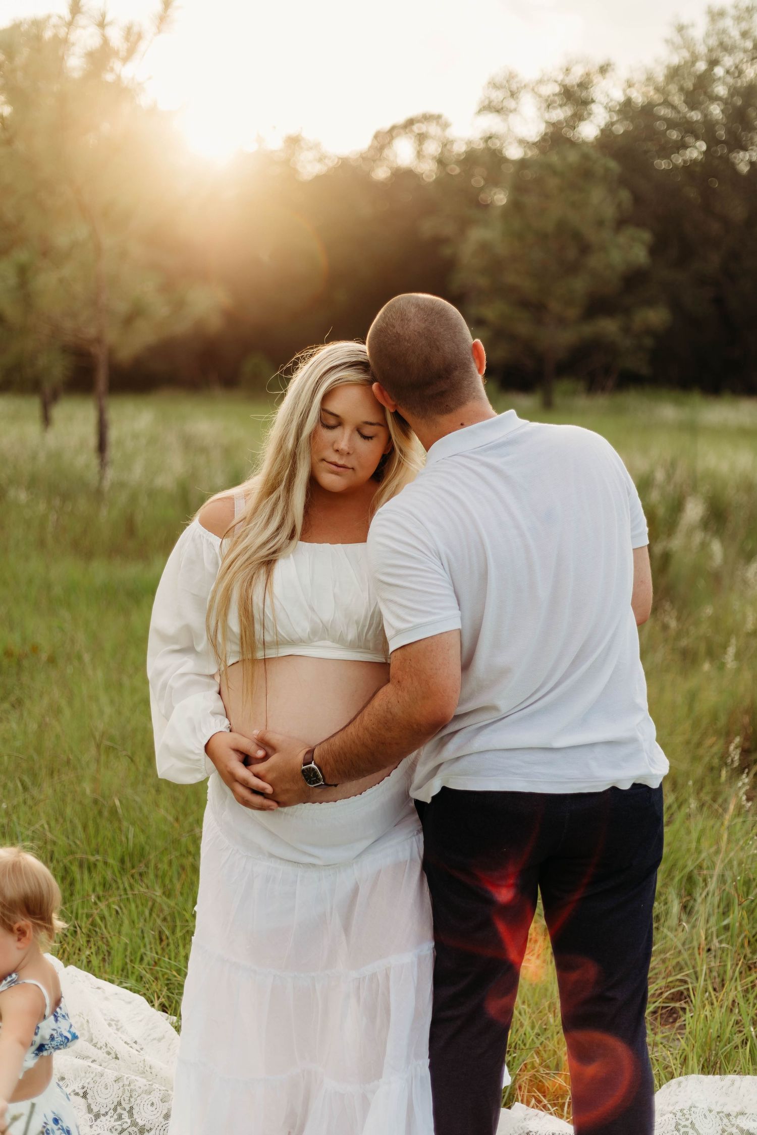 Couple tenderly embracing pregnant belly together in golden sunlit field at sunset.