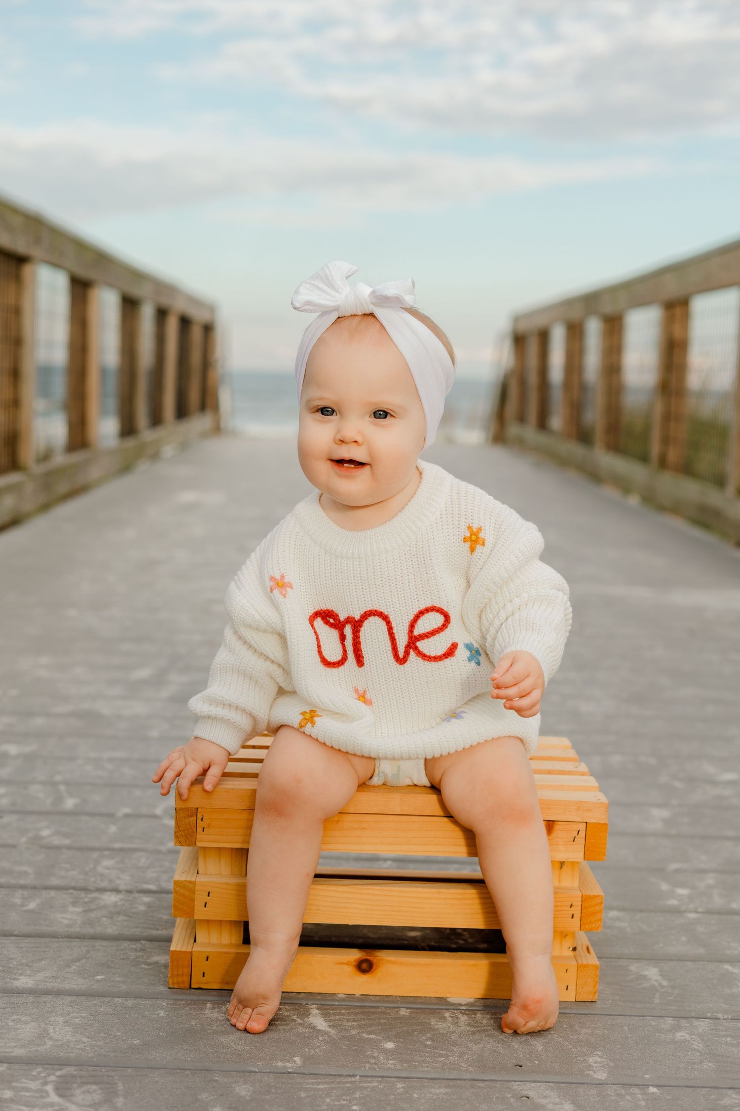 Baby girl in white 'one' sweater and bow headband sits on wooden crate on beach boardwalk for first birthday photos.