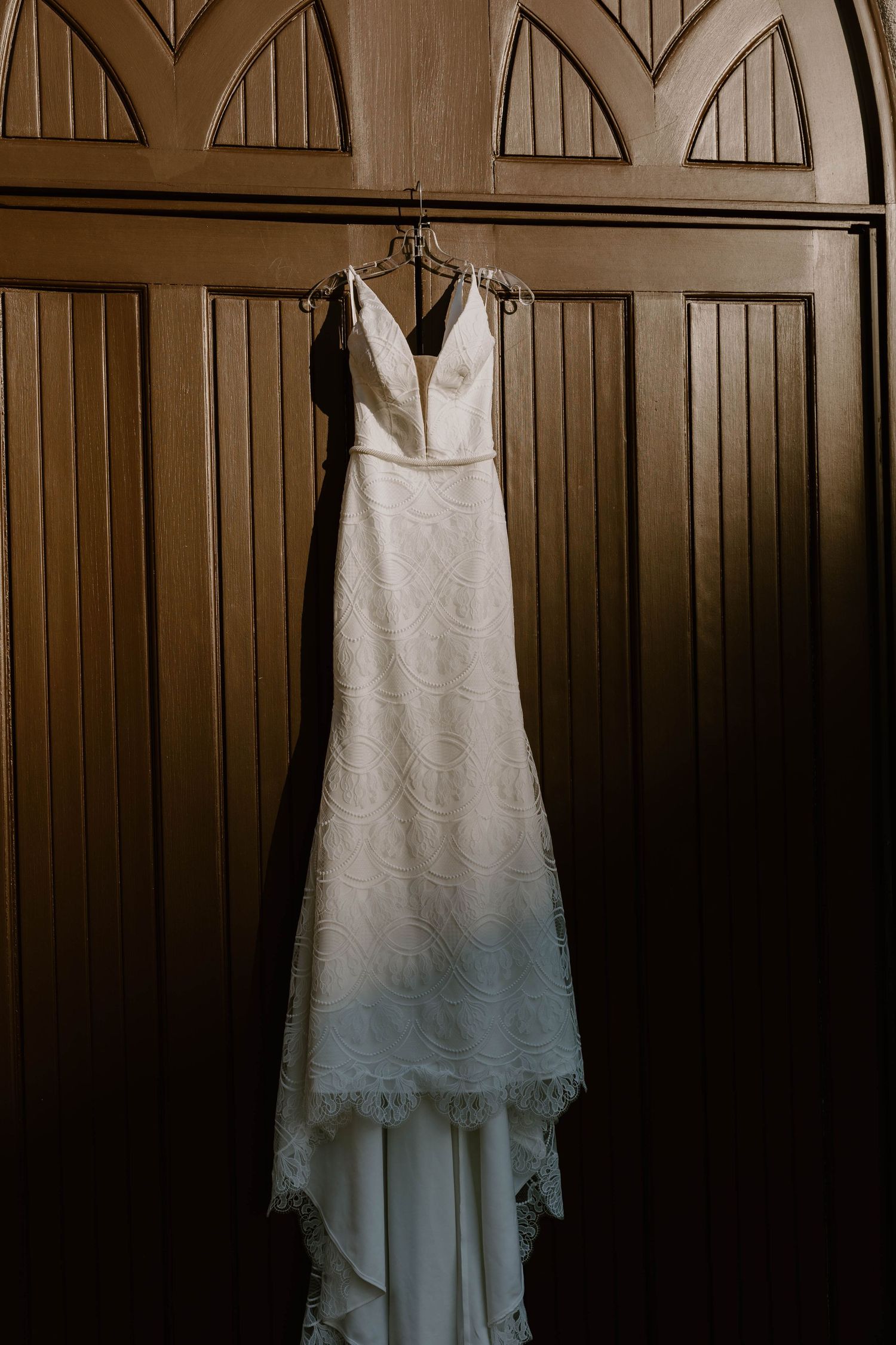 Sleek white lace wedding gown with V-neckline hangs on ornate wooden doors in dramatic moody lighting.