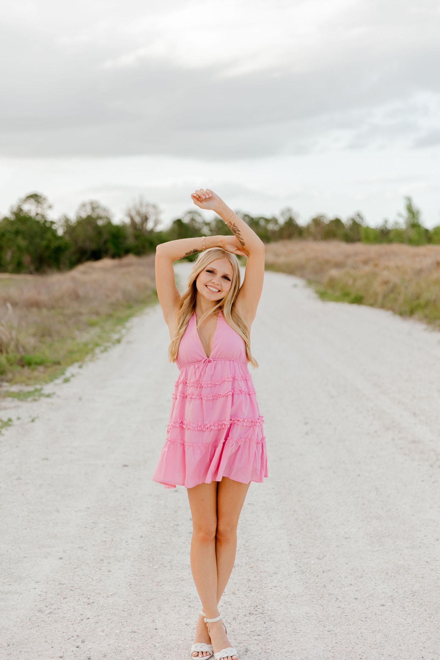 Blonde woman in pink mini dress posing on a rural dirt road with arms raised.