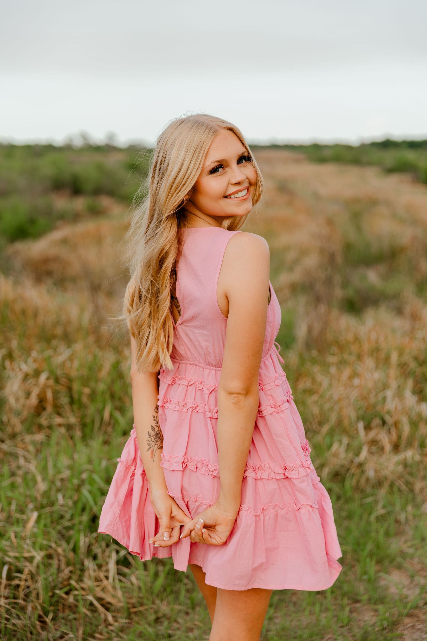Smiling blonde woman in pink ruffle dress posing in a golden field at sunset.