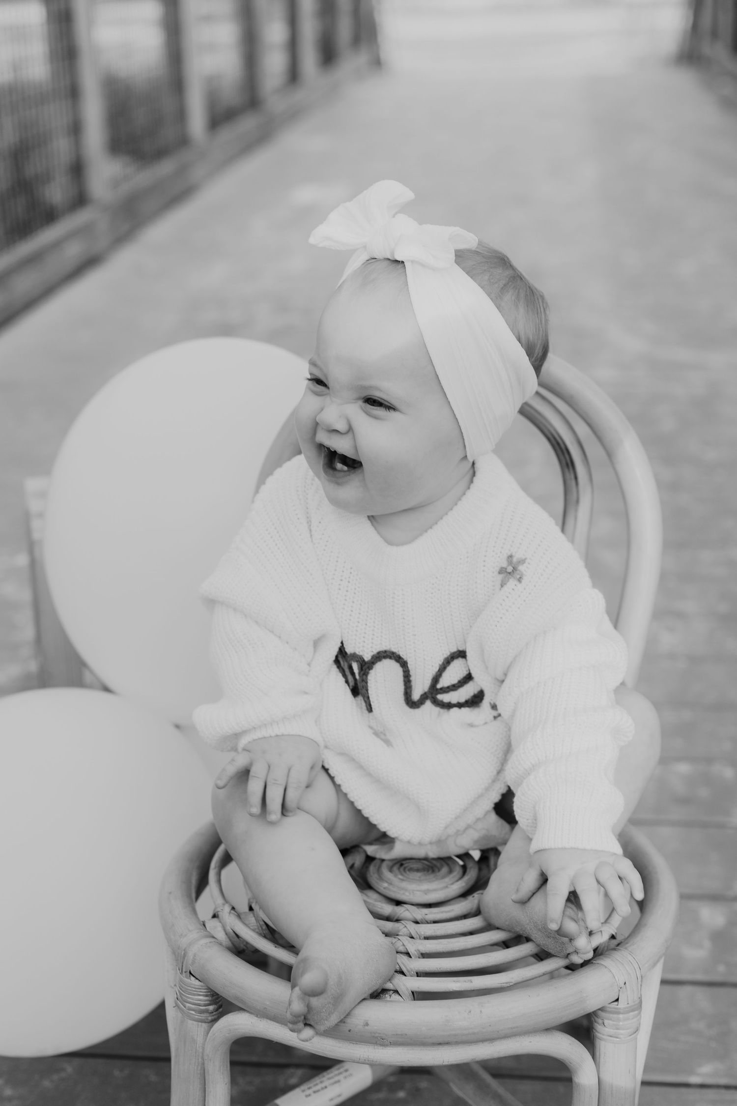 Laughing baby girl in white 'one' sweater and bow headband sits in rattan chair with balloons for birthday photos.