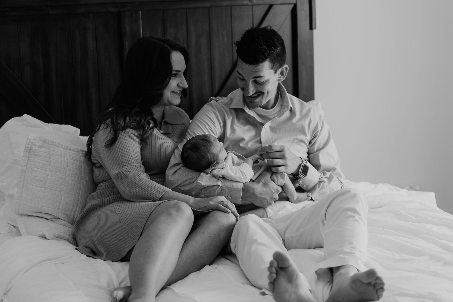 Black and white photo of happy parents sitting on bed smiling at their newborn.