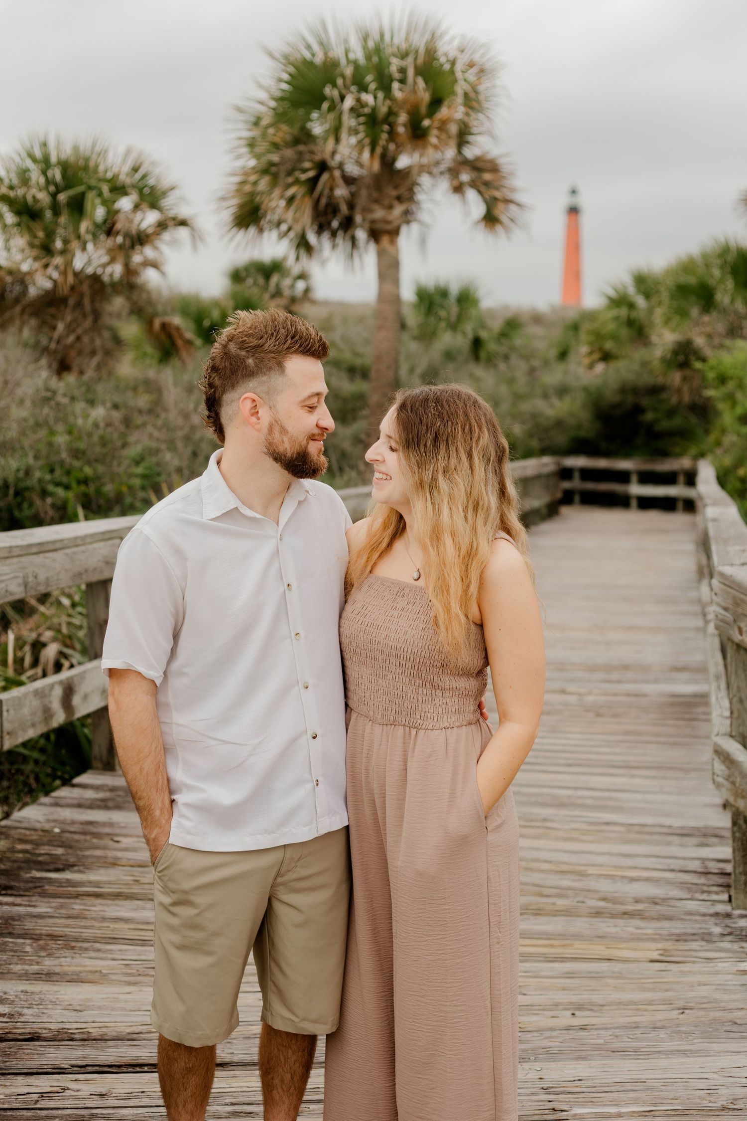 Couple smiling at each other on a beachside boardwalk with a lighthouse and palm trees in background.