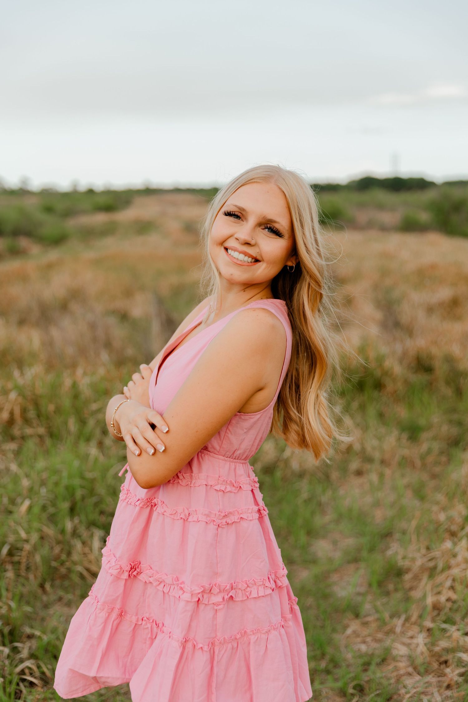 Blonde woman in pink ruffle dress smiling with arms crossed in a grassy field.