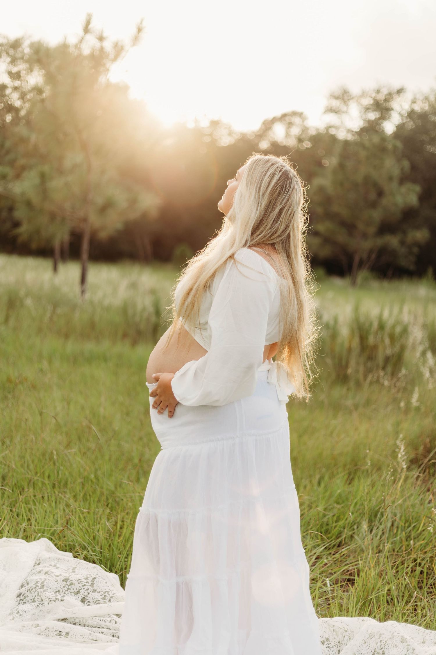Pregnant woman in flowing white dress cradles baby bump while standing in sunlit green meadow at golden hour.