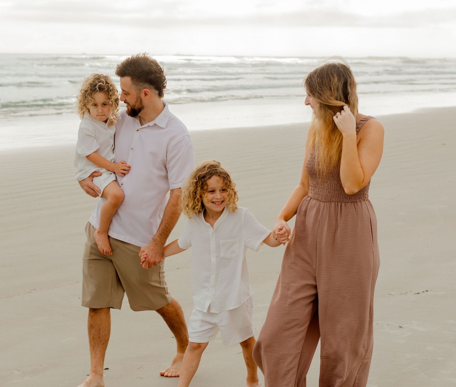 Family of four walking and playing together on a sandy beach with ocean waves in the background.
