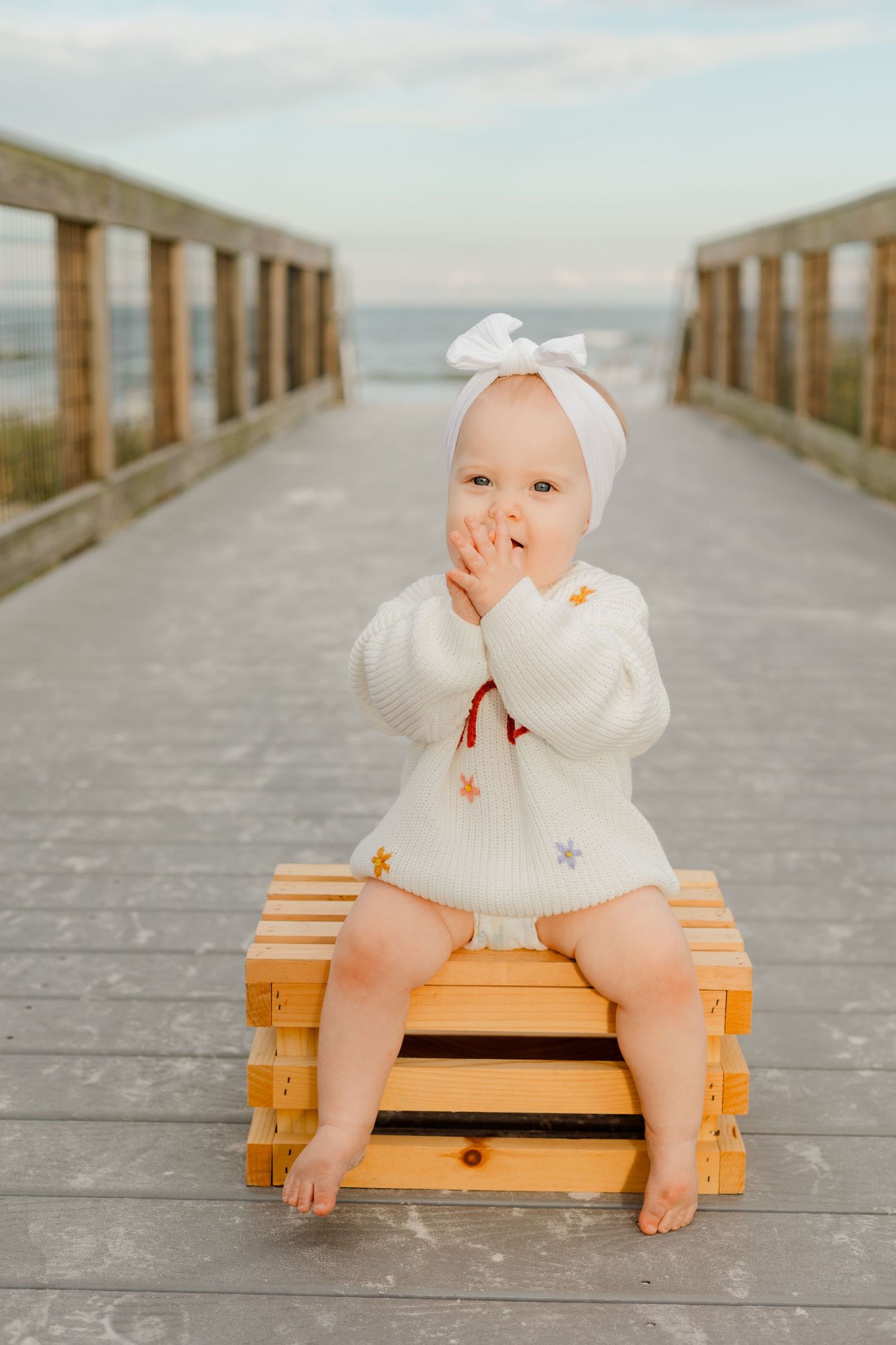 Adorable baby in white 'one' sweater sits on wooden crate on beach boardwalk, hands near mouth, first birthday.