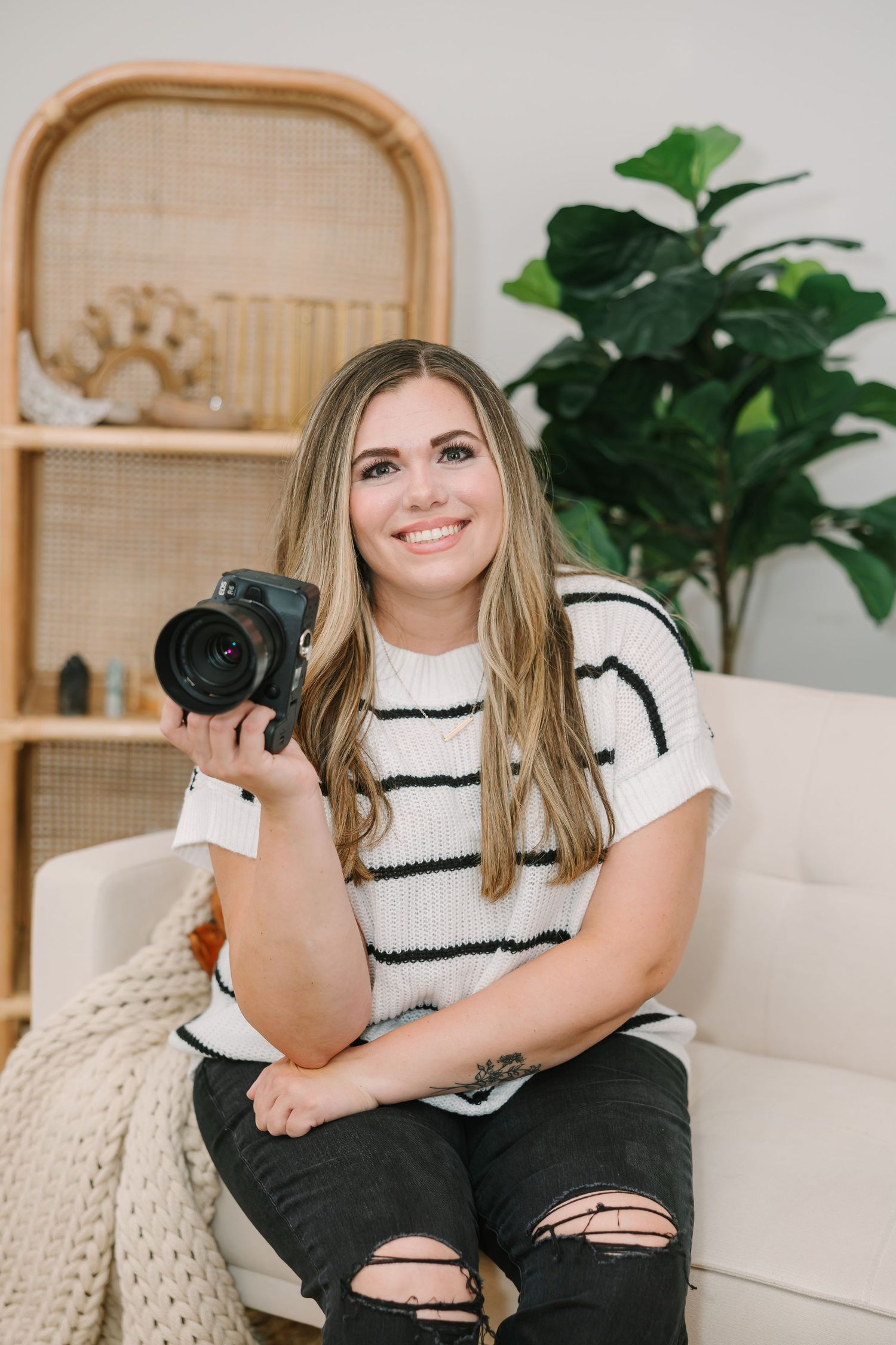 Smiling female photographer holding DSLR camera, seated on white couch with green plants and rattan decor behind her.