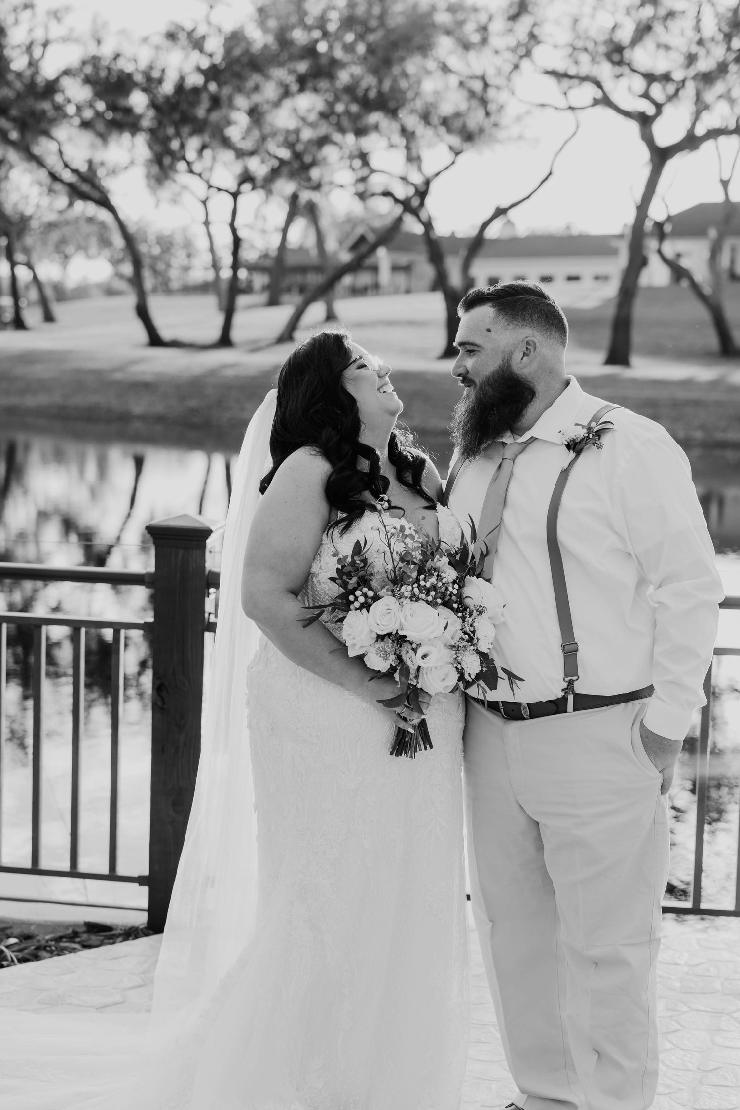Black and white photo of laughing bride and bearded groom in suspenders standing by a waterfront railing.