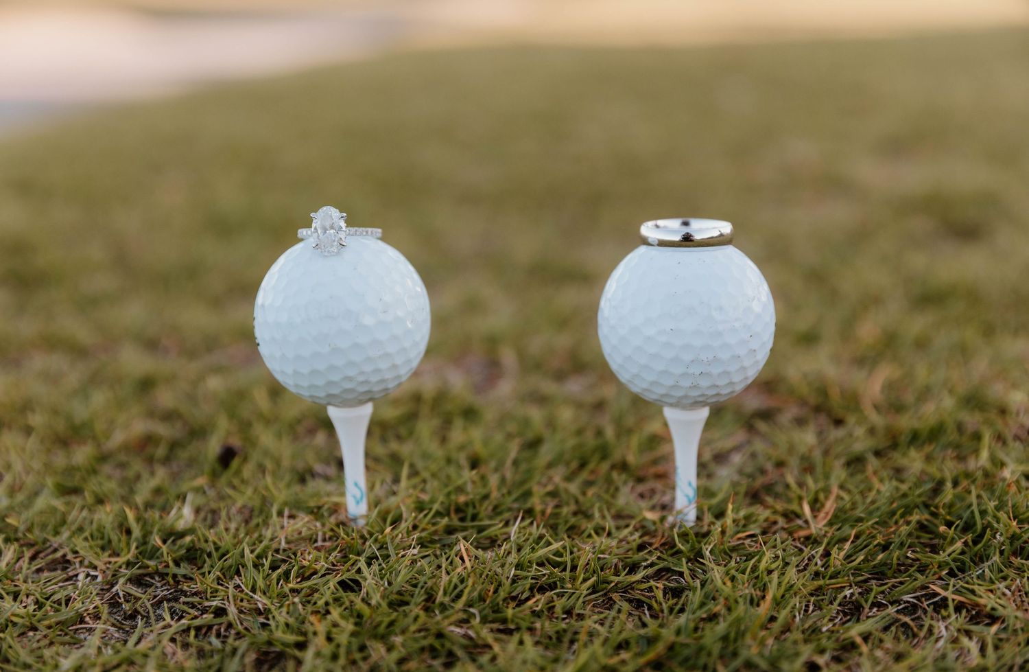 Two golf balls on tees in grass, each topped with a wedding ring, symbolizing a golf-themed wedding proposal.