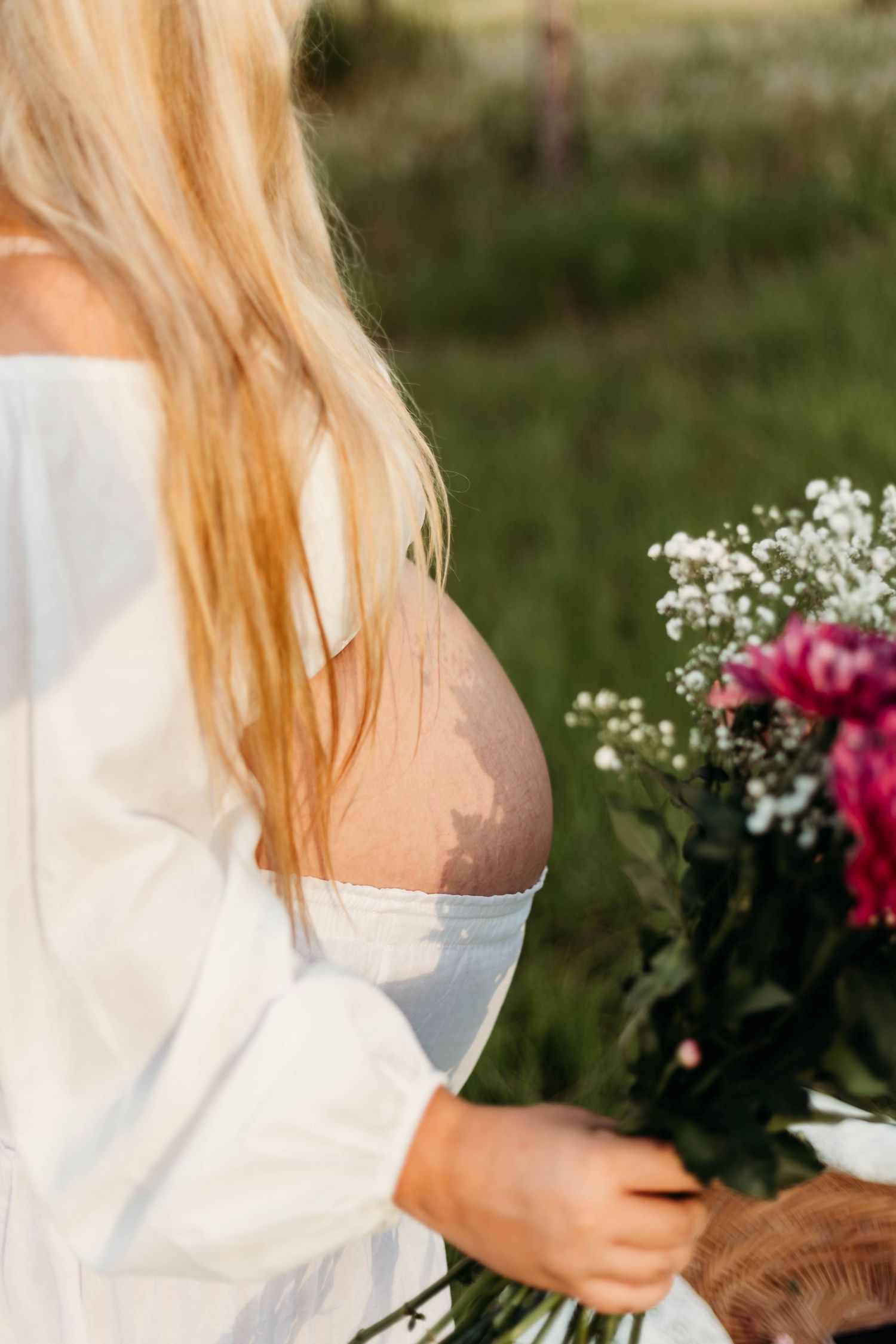 Side profile of pregnant woman in white off-shoulder dress holding colorful flower bouquet in green field.