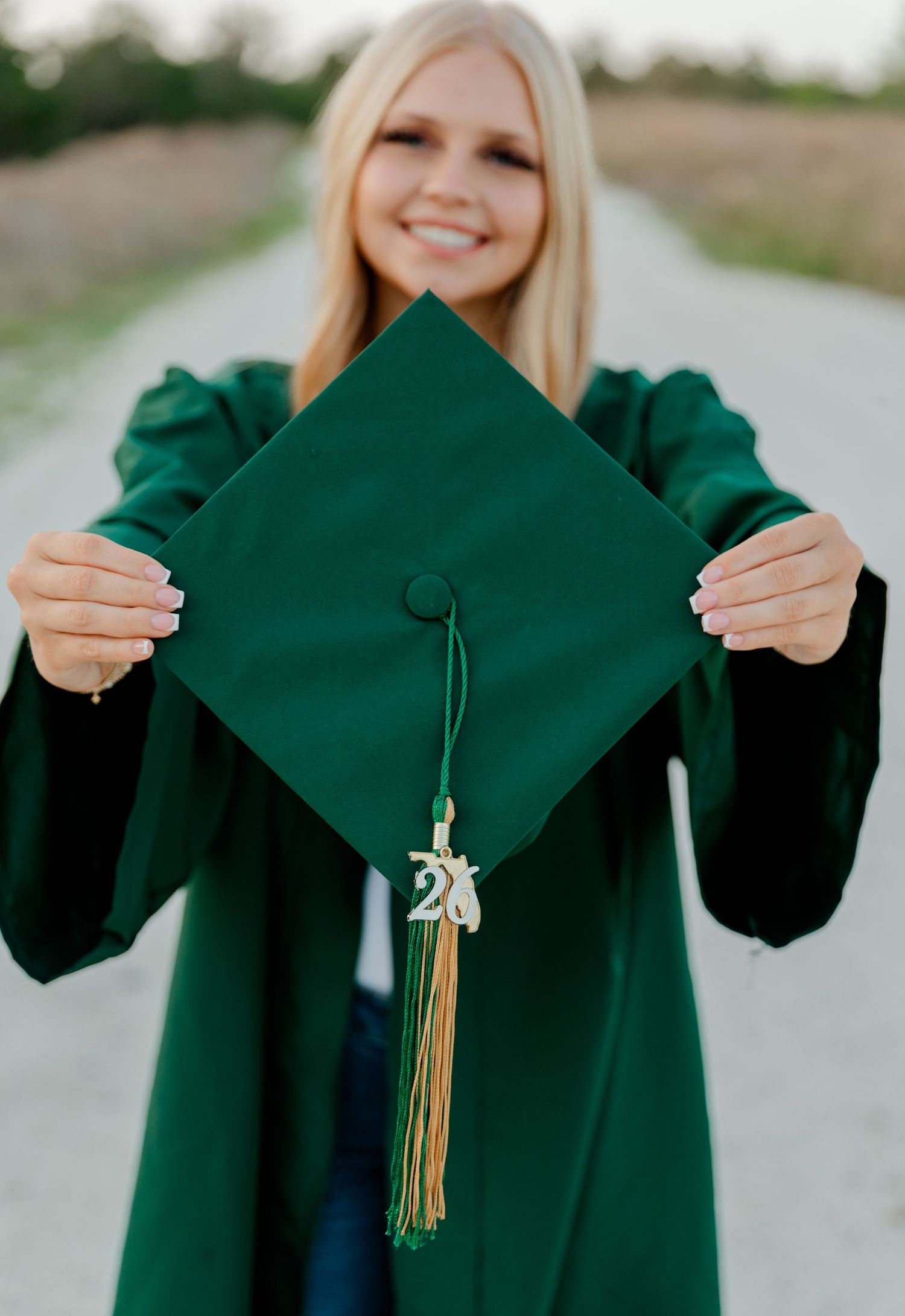 Smiling blonde graduate holding green cap and gown toward camera on a dirt road.