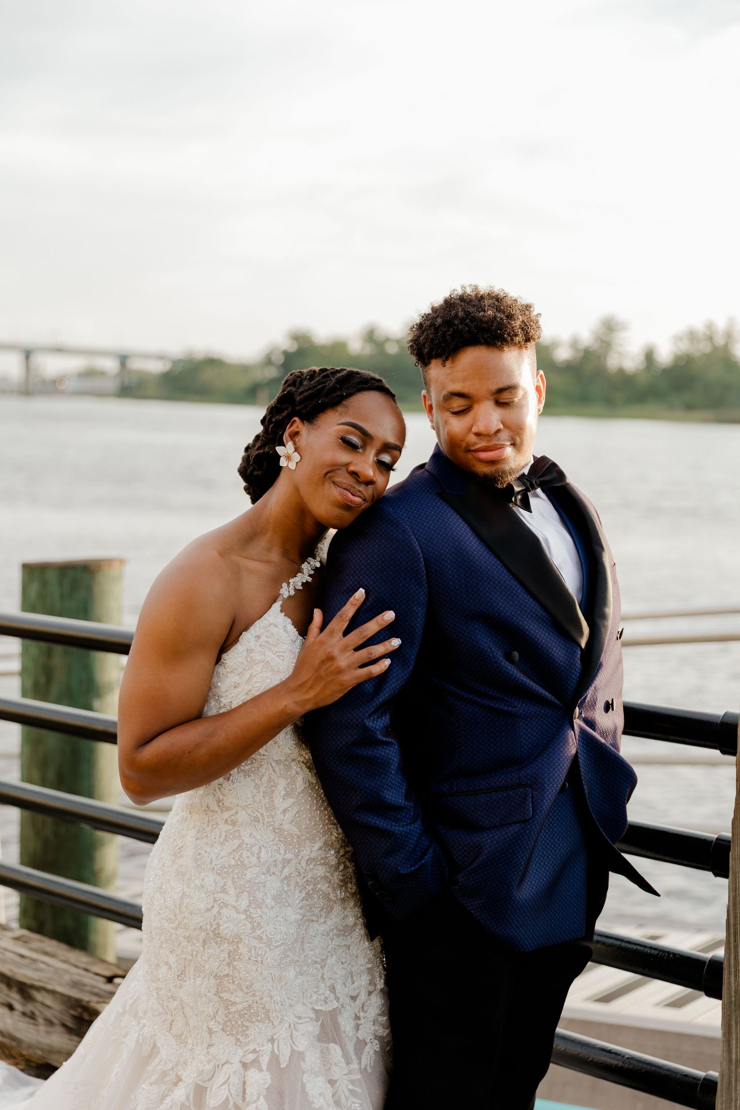 Black bride in lace wedding gown and groom in navy tuxedo embracing on waterfront dock at sunset.