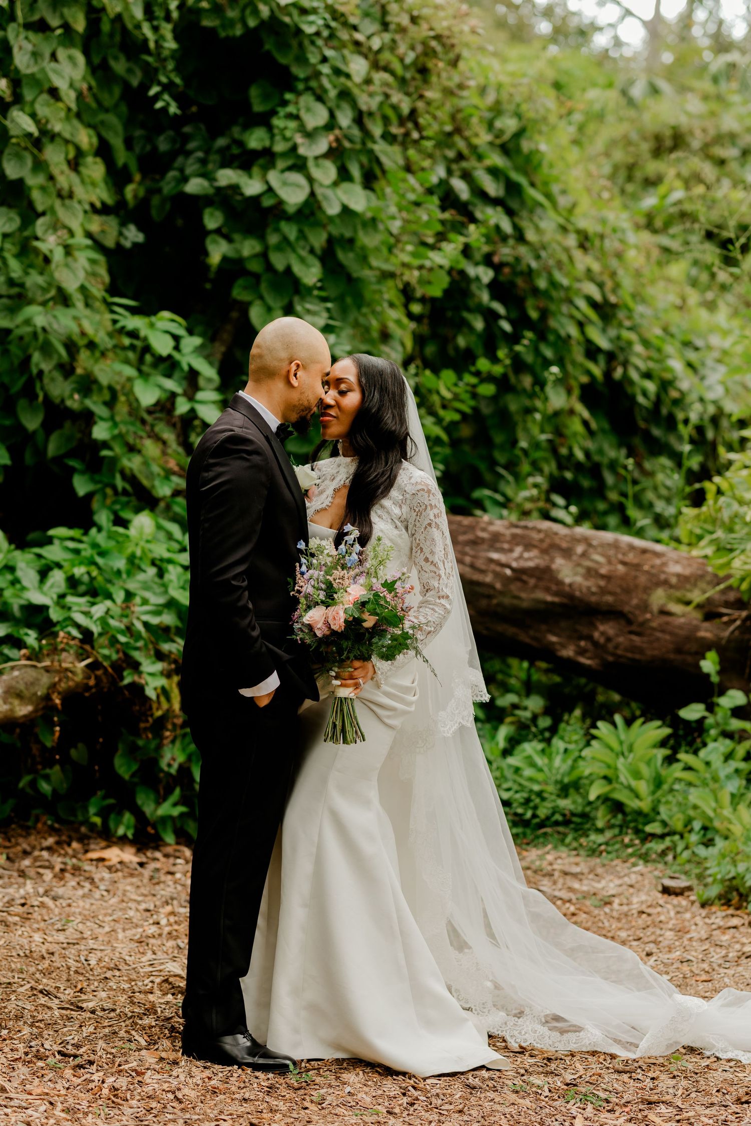 Groom kissing bride on the forehead in a woodland setting, bride wearing a lace veil and holding a colorful bouquet.