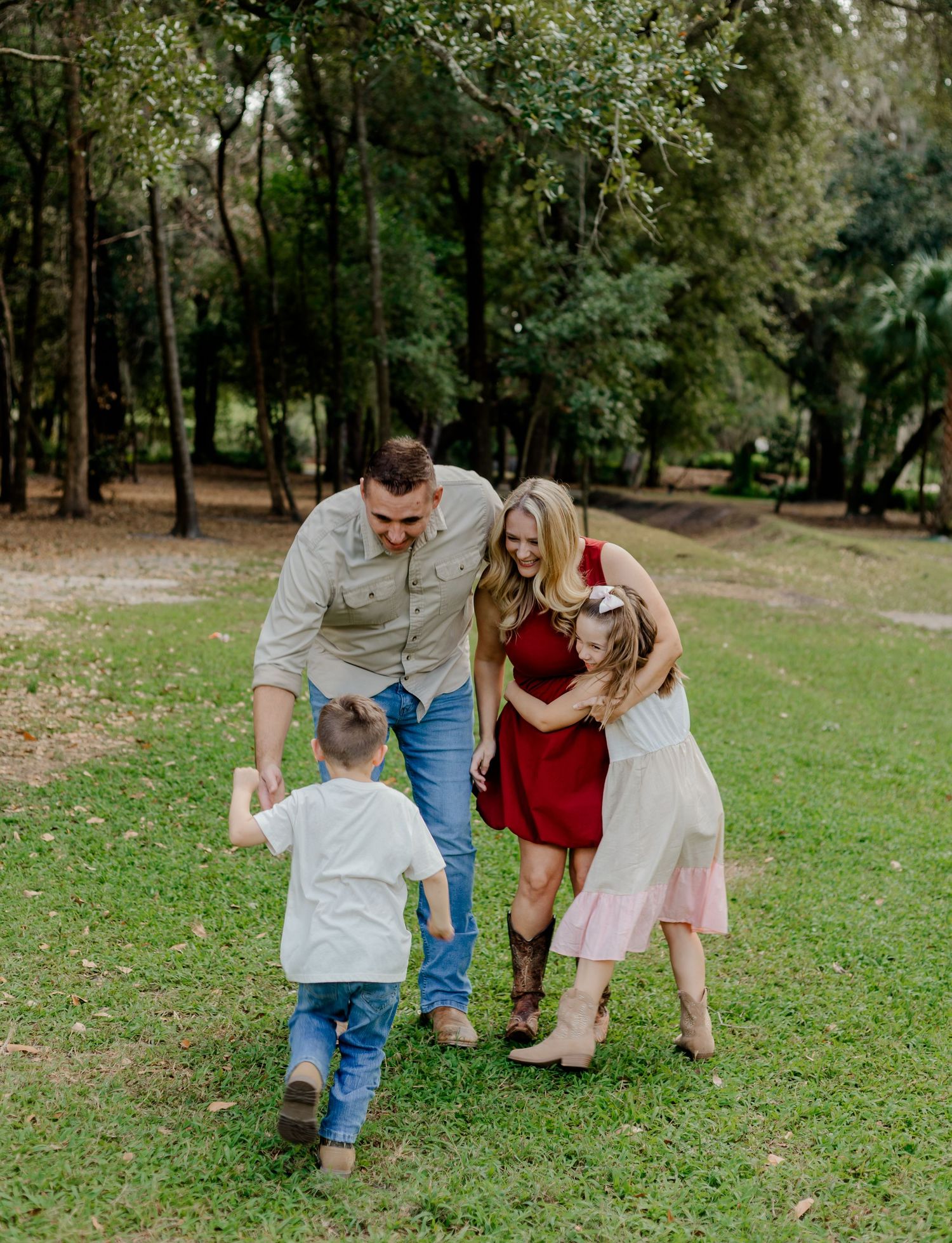 Candid family photo of parents and two children playing together on a green lawn surrounded by trees.