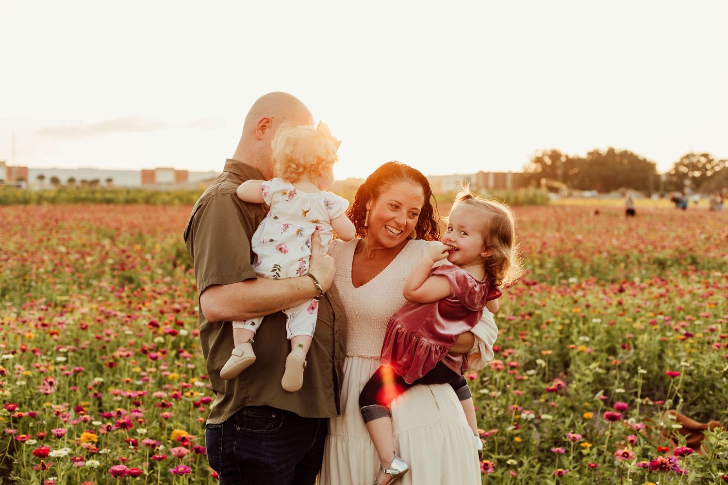 Happy family with two toddler girls laughing together in a vibrant flower field at golden sunset.