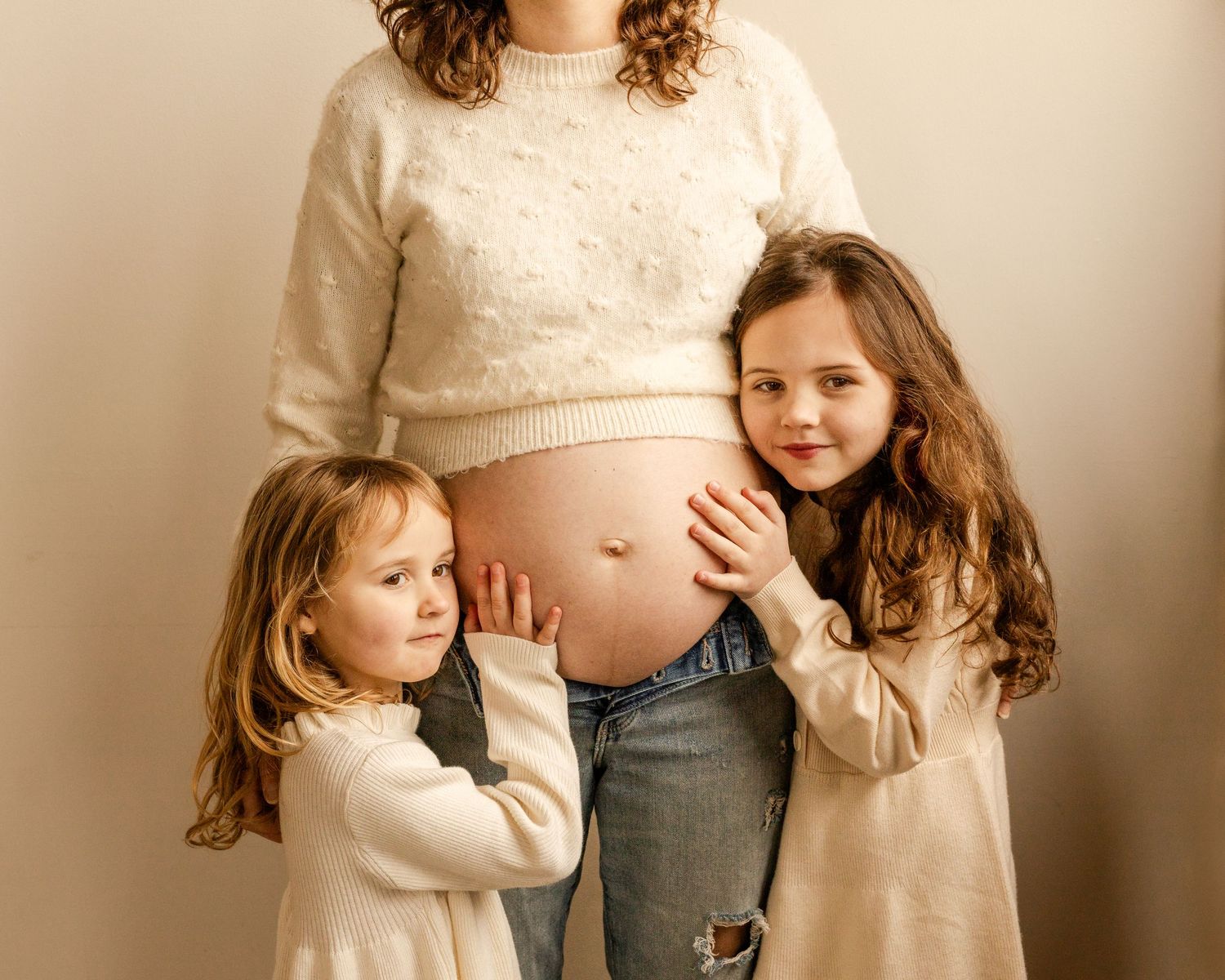 Children in cream-colored sweaters touch a pregnant belly wearing ripped jeans against neutral background. Studio Family Maternity session