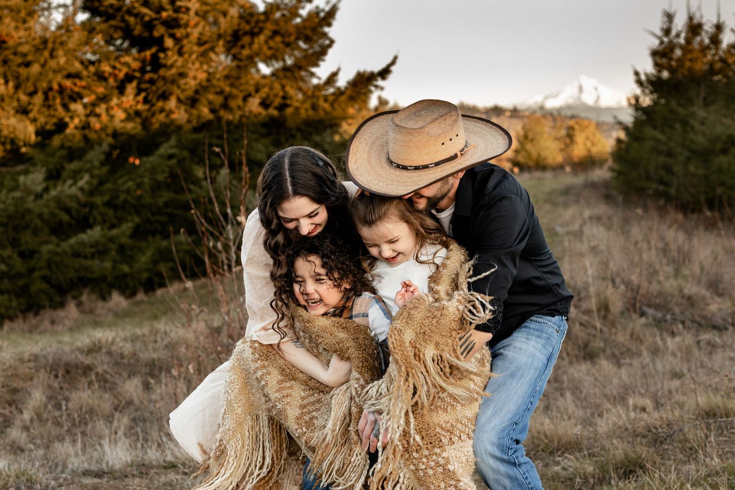 A family enjoys a playful moment with their two fluffy golden retrievers in an autumn field at sunset.