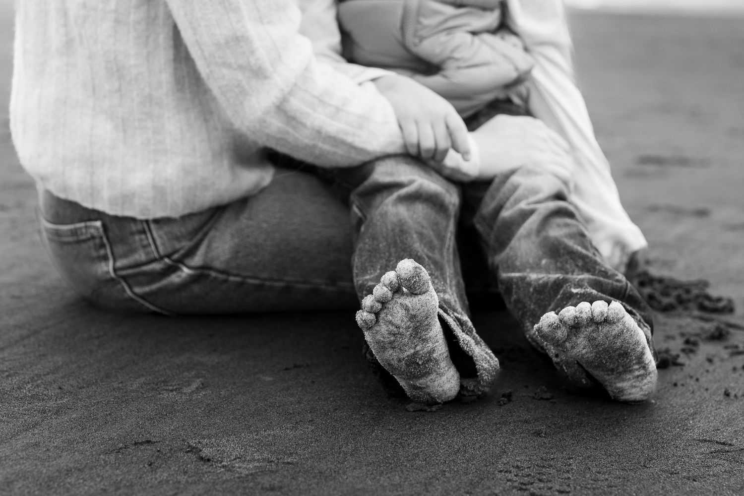 Black and white photo of a parent and child sitting on sandy beach, focusing on their bare sandy feet.