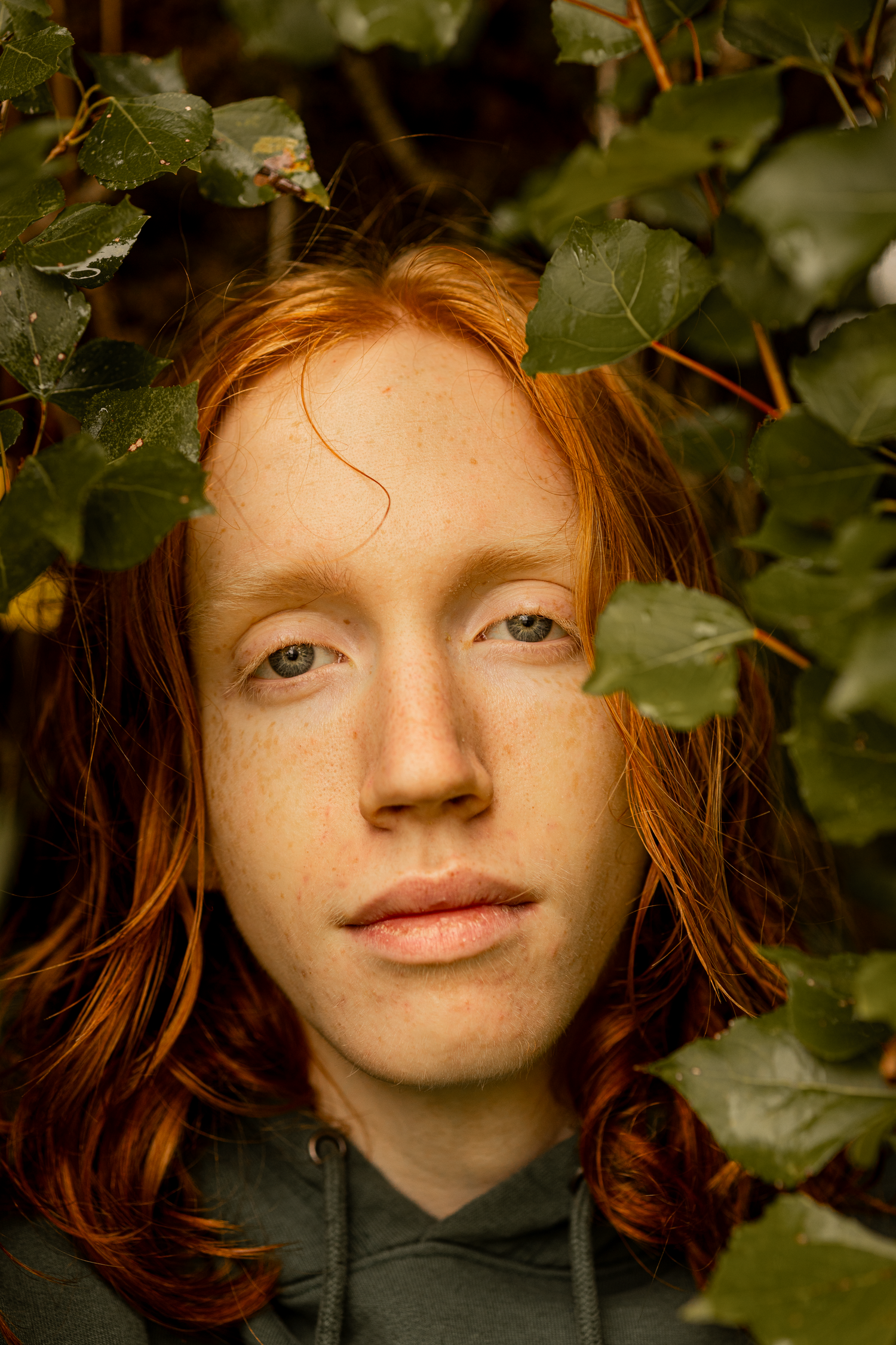 Portrait of person with red hair framed by green leaves in natural lighting.
