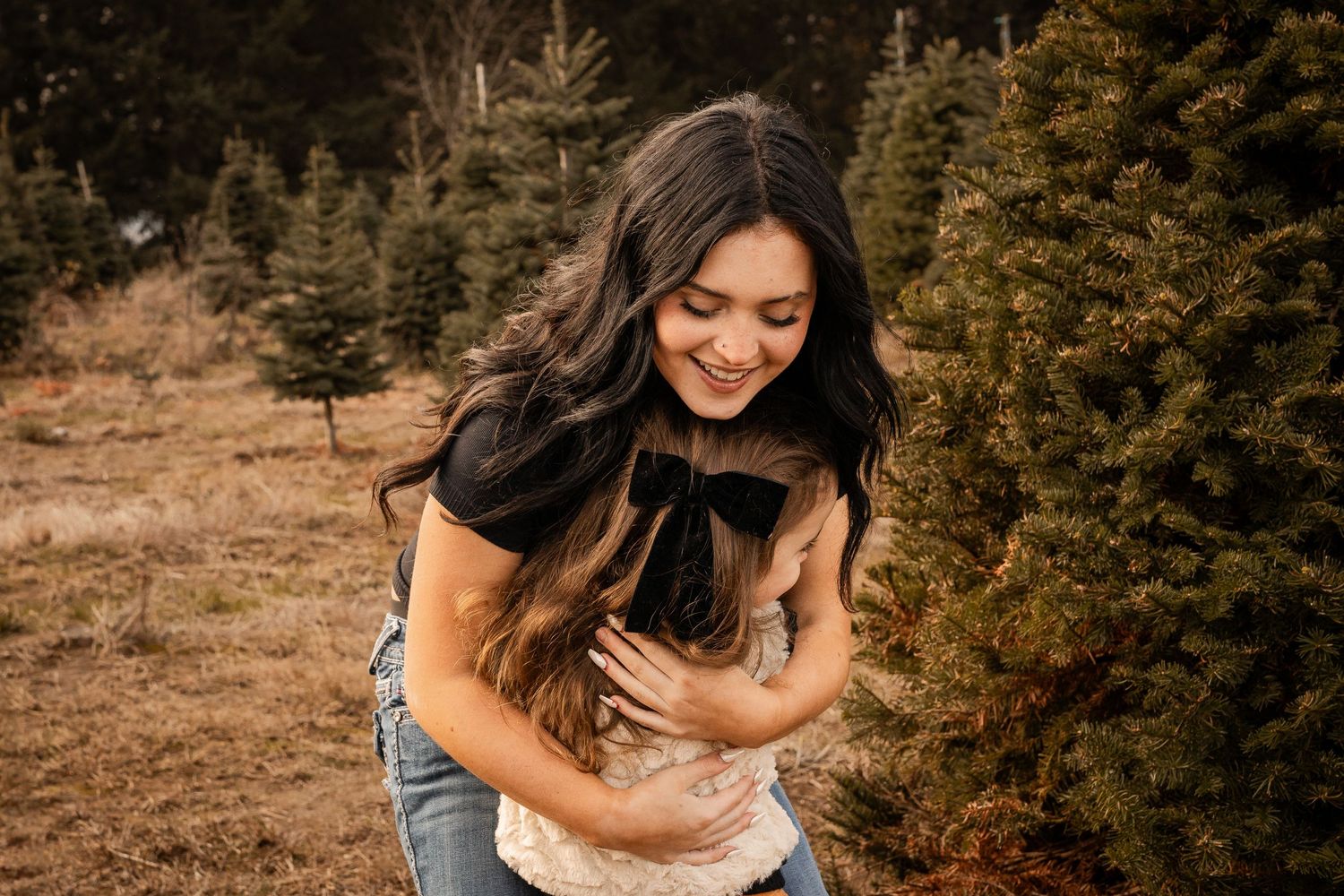 Woman smiling while holding and petting a cat outdoors near evergreen trees.