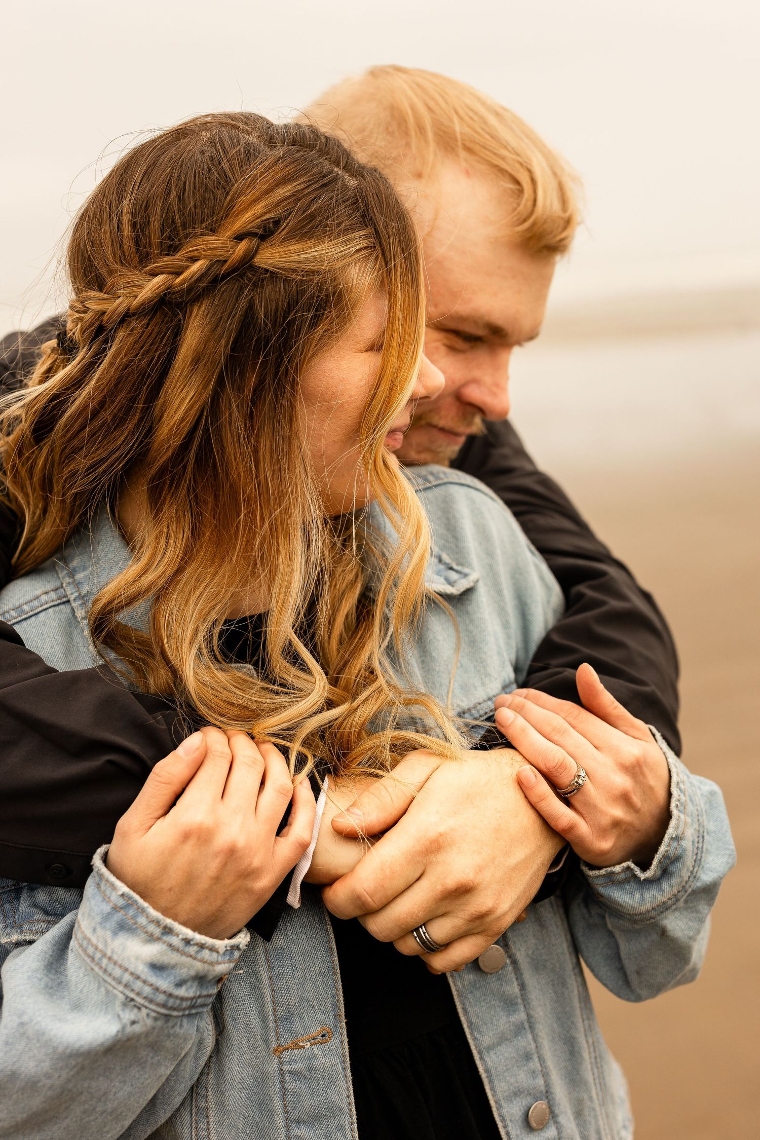 A couple embraces warmly on a misty beach, the woman in a denim jacket smiling as the man wraps his arms around her.