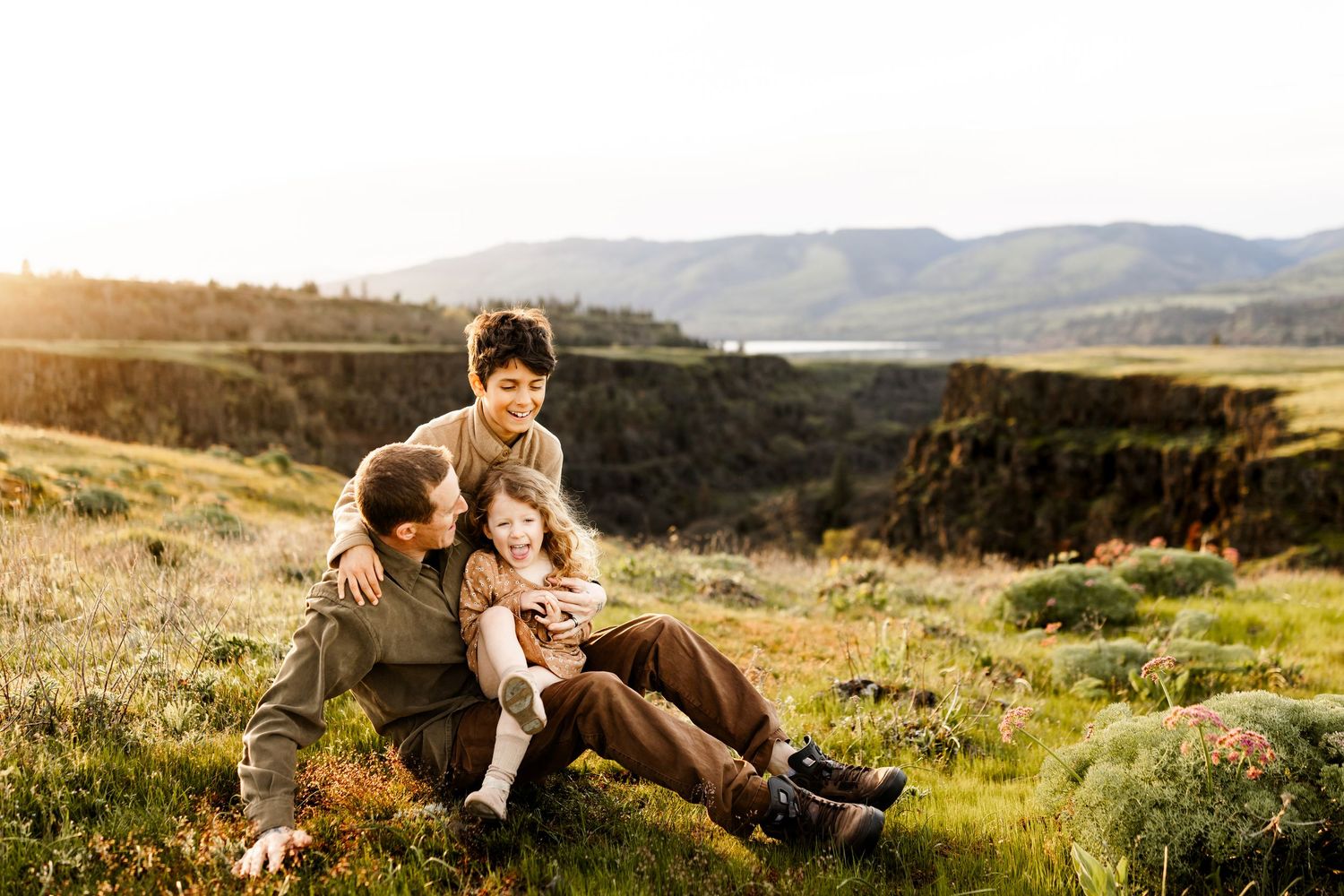 Family of three sitting together on a grassy hillside with scenic mountain and lake views in warm golden light.