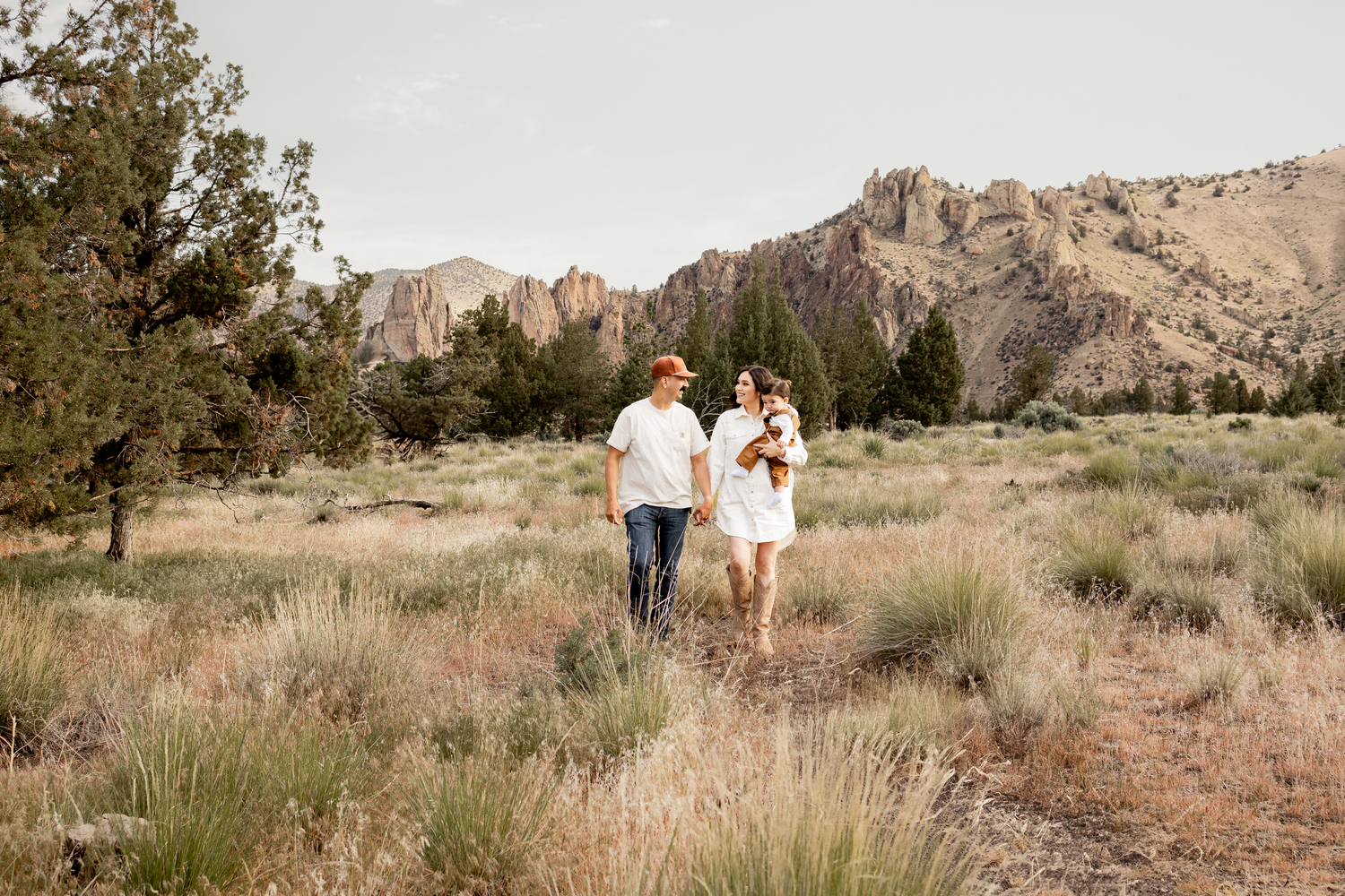 Couple walking hand-in-hand through a desert trail with rocky mountain peaks and sagebrush at Smith Rock State Park.