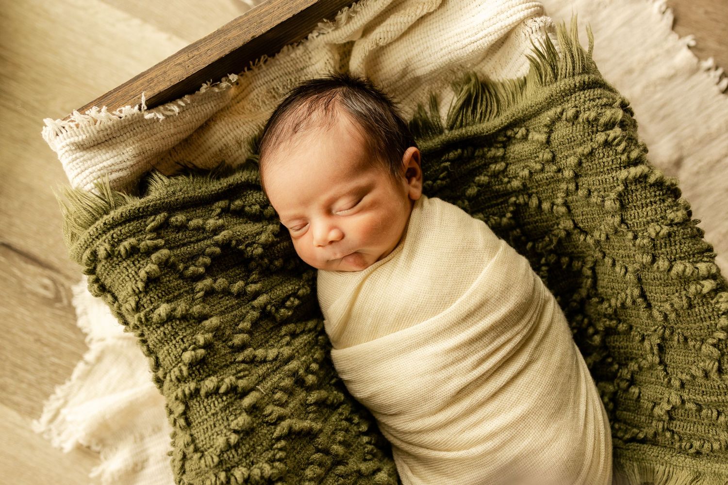 Newborn baby swaddled in cream wrap sleeping peacefully on green textured pillow in professional photography setup.
