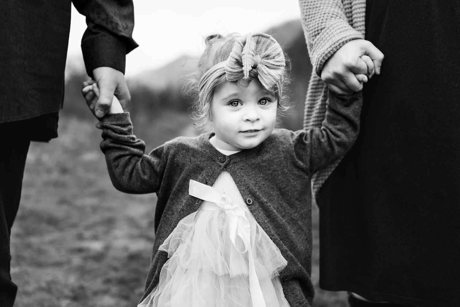 Black and white photo of a toddler girl in tutu dress holding hands with two adults outdoors.