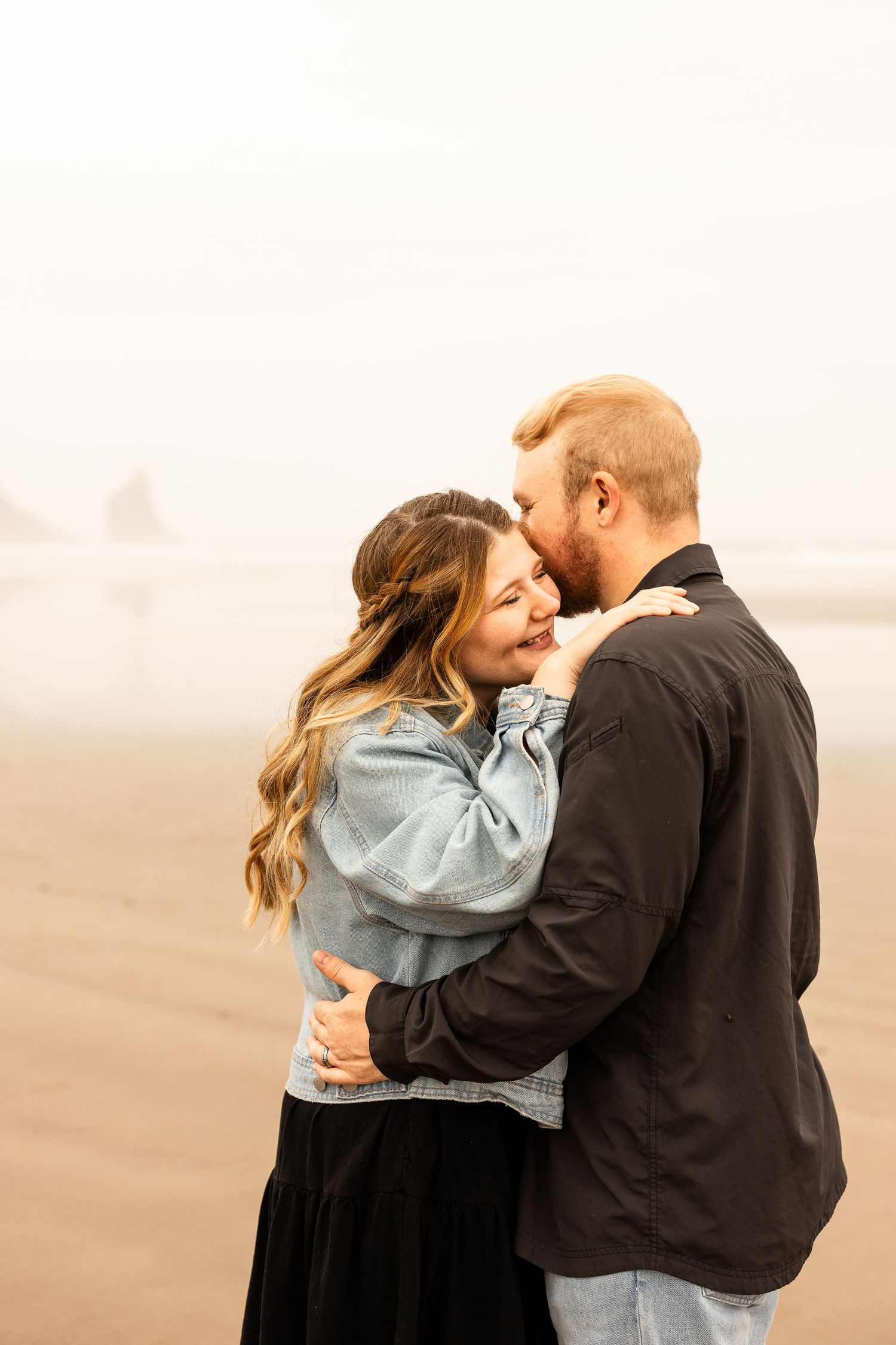 Couple embracing and kissing on a misty beach, woman in denim jacket smiling joyfully.