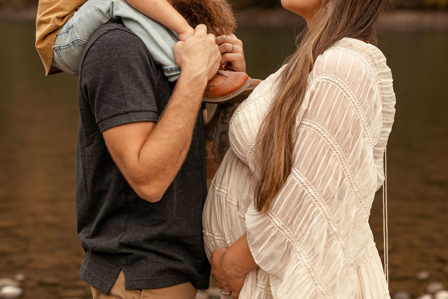 Pregnant woman in white lace dress with partner holding child near water during maternity photo session.