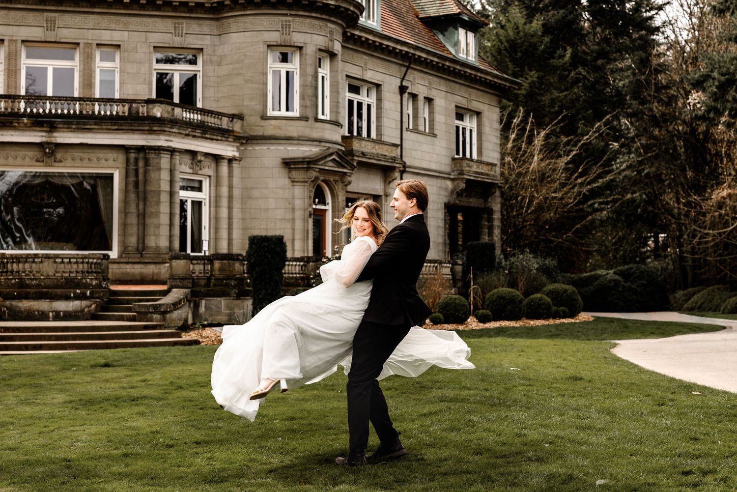Groom lifting bride in white gown outside elegant stone mansion during romantic outdoor wedding photo session.