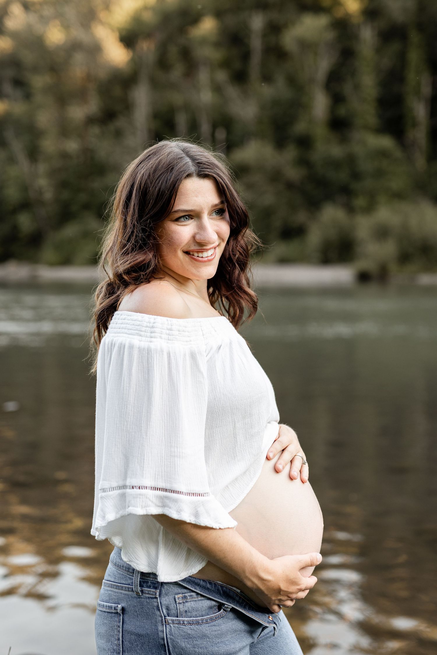 Smiling pregnant woman in white off-shoulder top and jeans cradling her baby bump outdoors near a serene river.