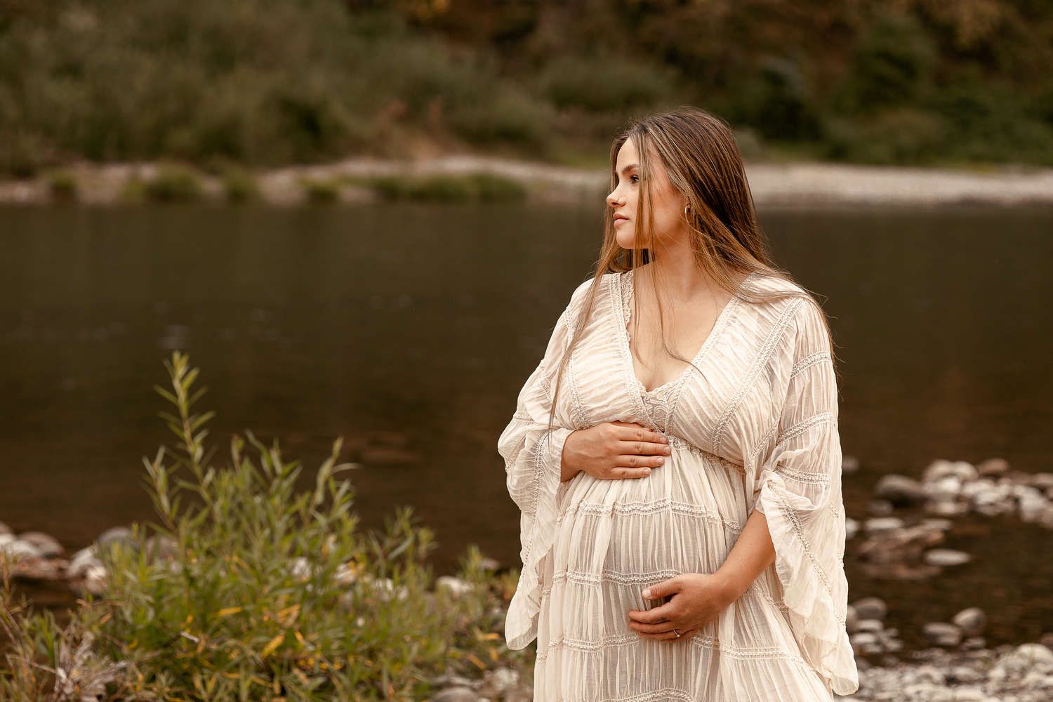 Pregnant woman in white lace dress cradling baby bump by a serene river surrounded by lush greenery.