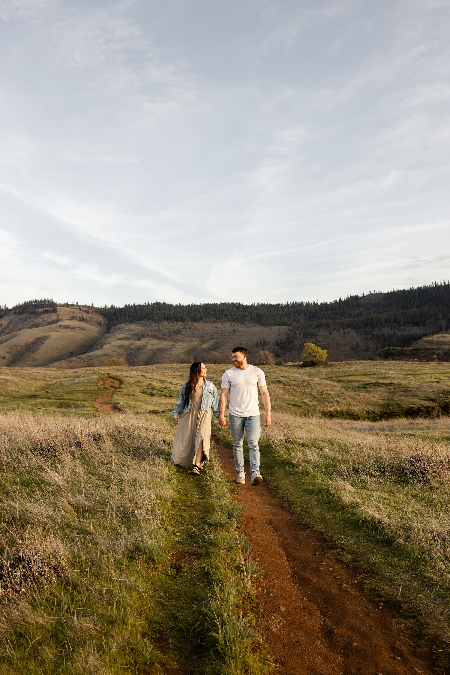 Couple holding hands walking on a dirt path through green meadows with rolling hills in the background.