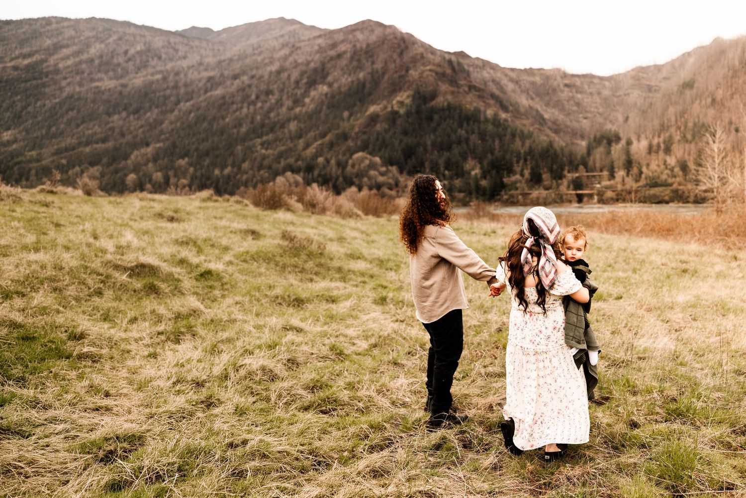 A mother and two children hold hands in a grassy mountain meadow during a warm autumn day outdoors.