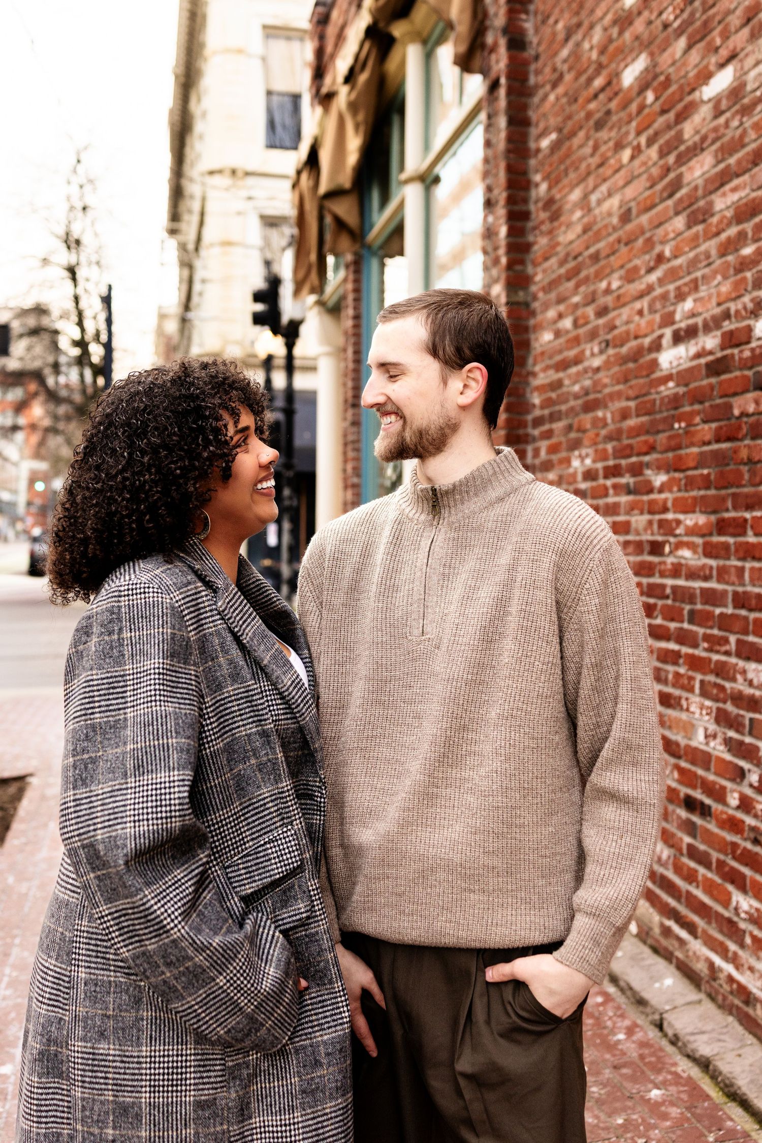A smiling interracial couple standing together on a brick-lined urban street, dressed in stylish fall outerwear.
