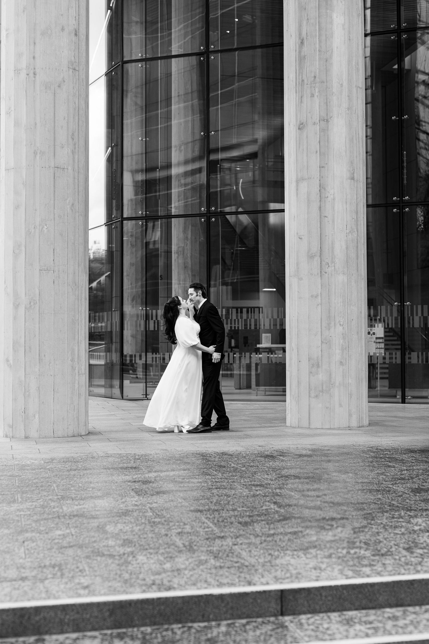 A couple in wedding attire embrace under modernist concrete columns with glass walls in a black and white photograph.