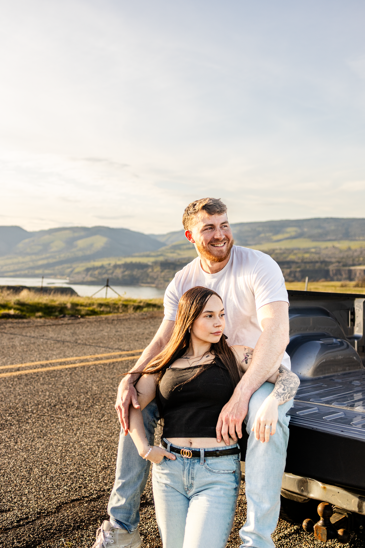 A smiling couple leans against a pickup truck with mountain lake views, wearing casual jeans and t-shirts on a sunny day.