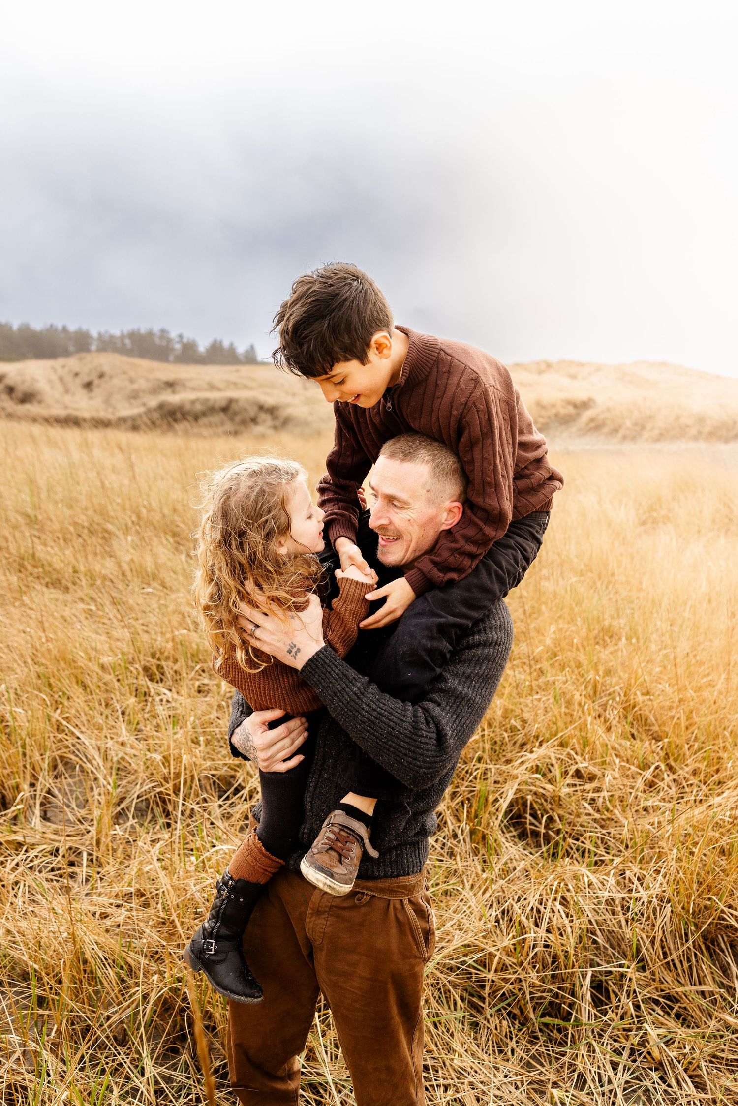 A father joyfully holds his two children in a golden autumn field under a cloudy sky.
