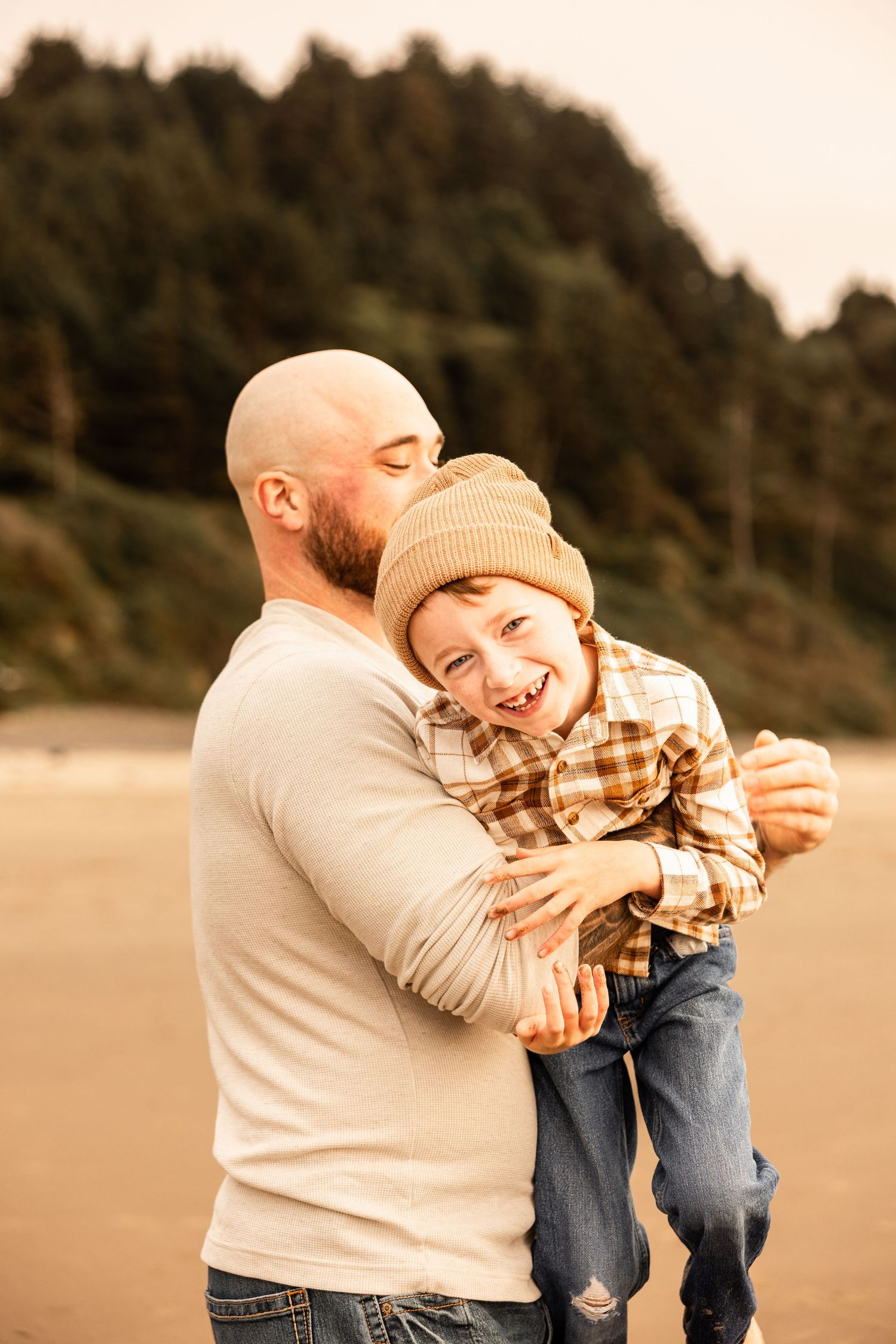 A bald father hugs his laughing child on a beach, both wearing casual fall clothing with trees in the background.