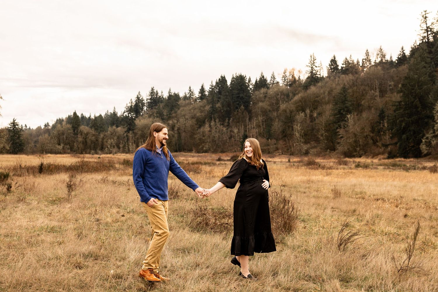 Couple holding hands in golden field with evergreen forest backdrop during fall outdoor engagement photo session.