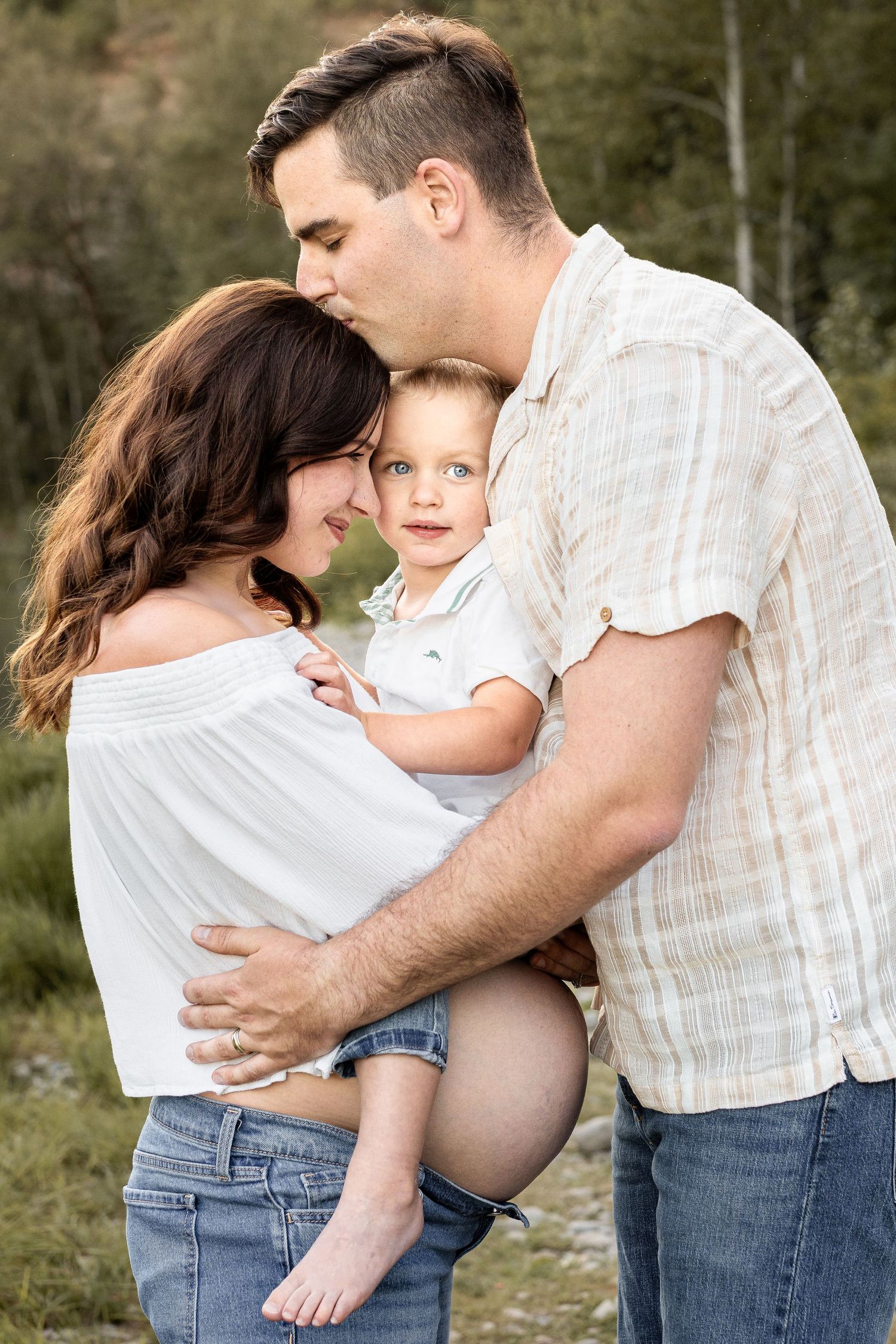 A loving family portrait of parents holding their toddler son outdoors, with dad kissing mom's head.