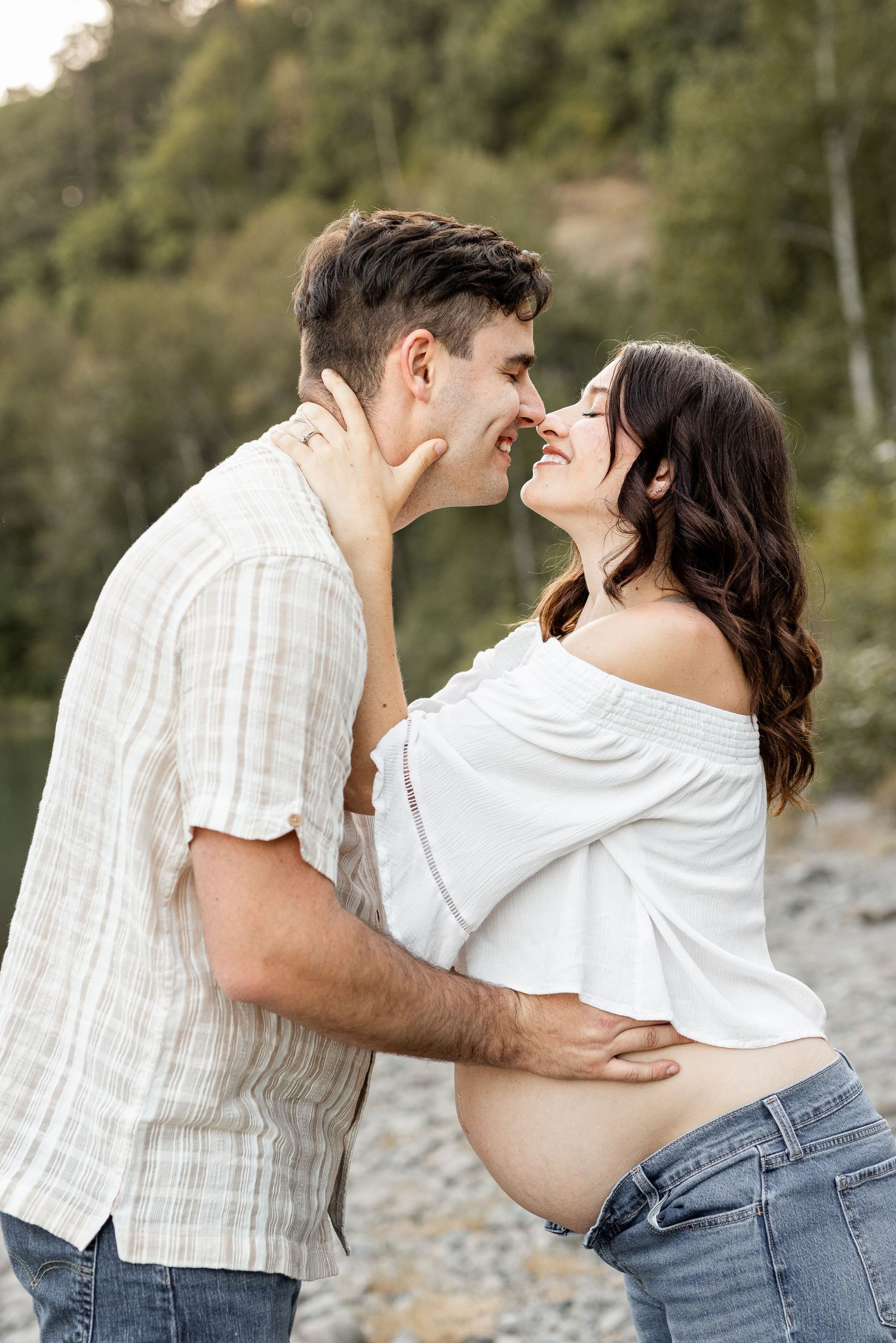 Couple sharing a tender moment, pregnant woman in white off-shoulder top and jeans, outdoors with greenery background.