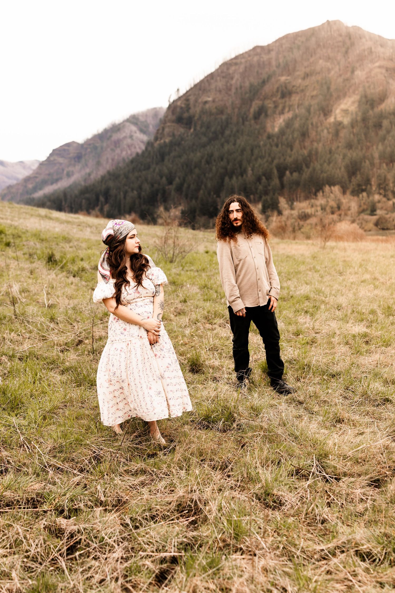 A couple stands in a mountain meadow, woman in floral dress with headscarf, man with long hair in casual attire.