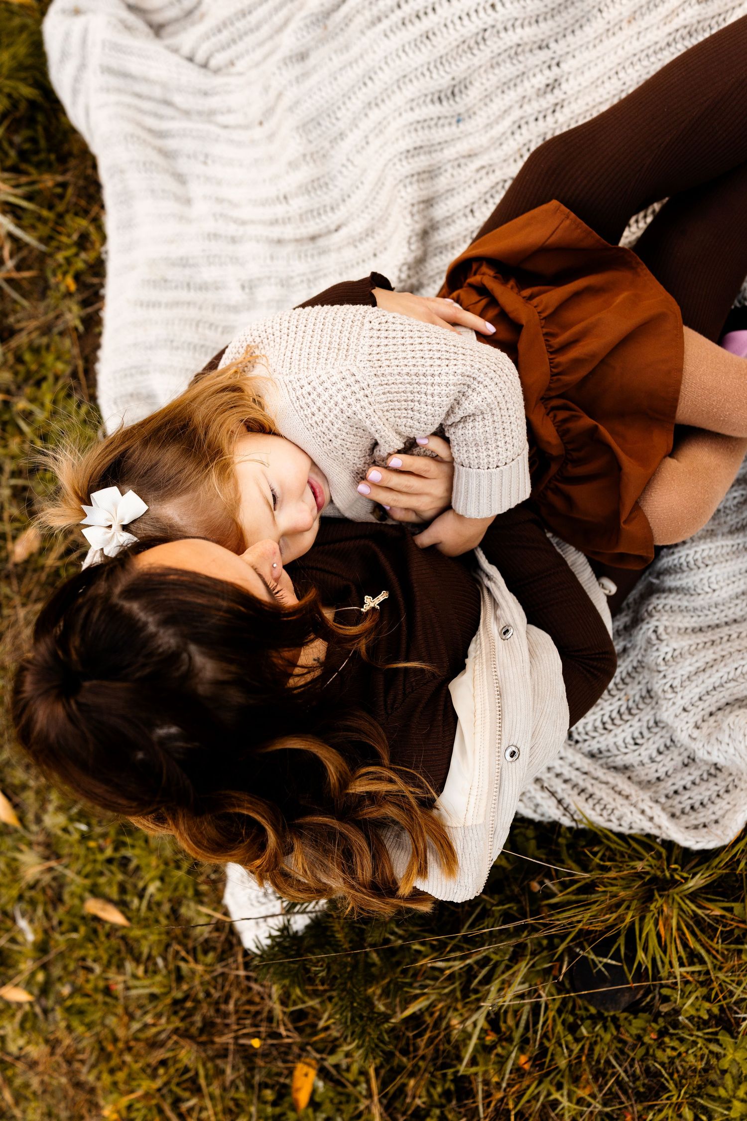 Mother and daughter lying on a white knit blanket on grass, sharing a tender moment in autumn.