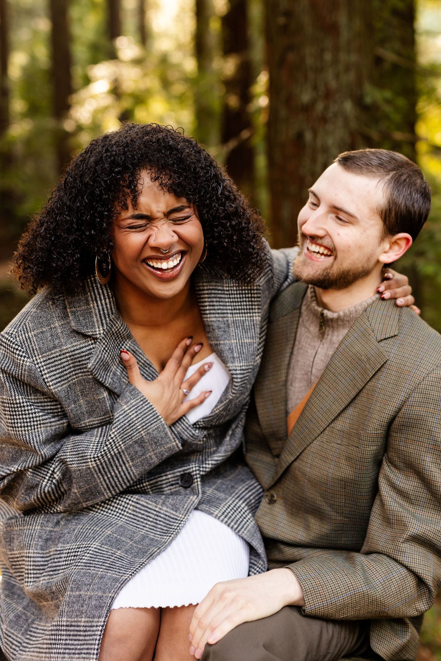 A laughing interracial couple embracing in a forest, wearing elegant autumn outfits in gray and olive tones.