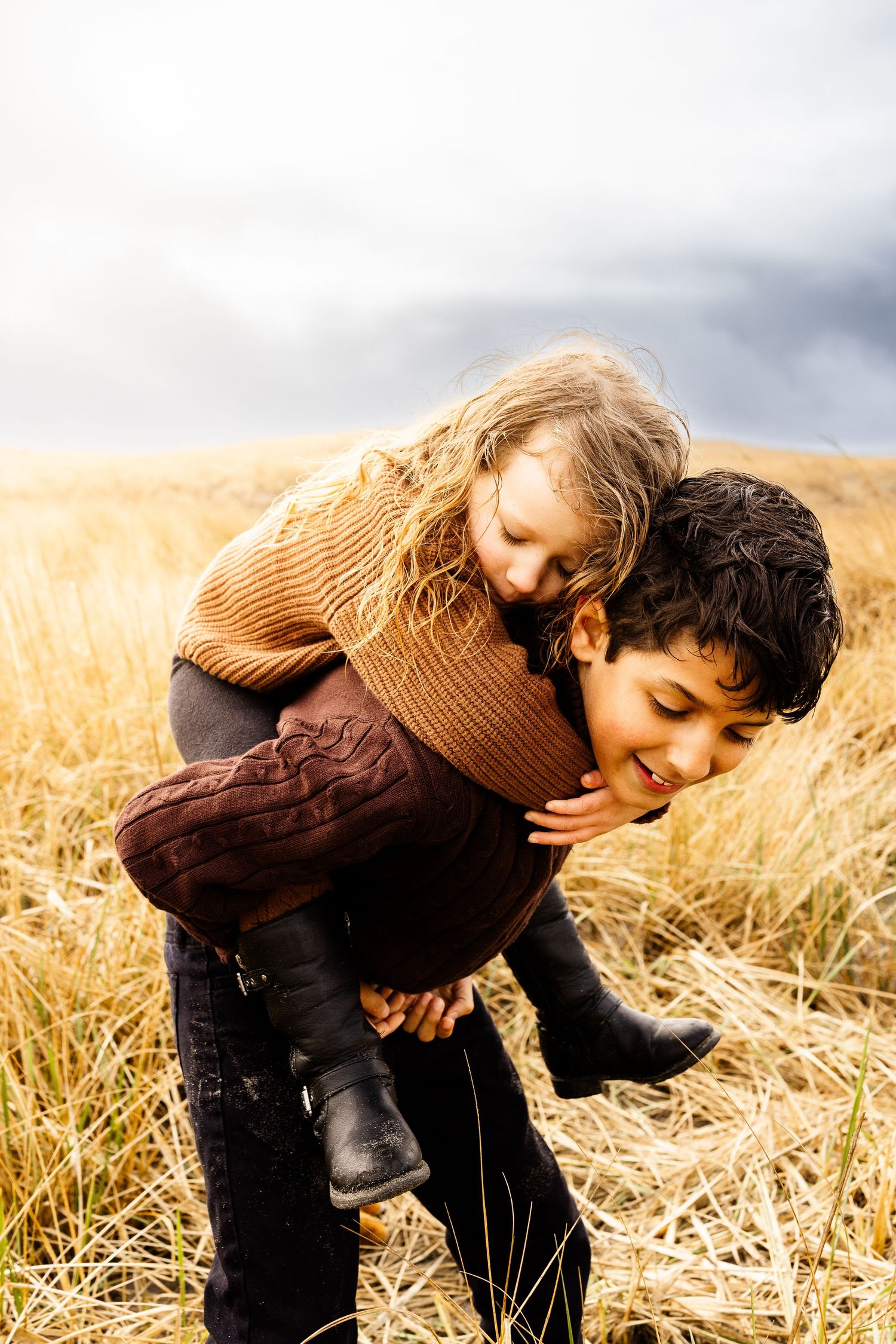 Two children playing piggyback in a golden wheat field under a cloudy sky, wearing cozy autumn sweaters.